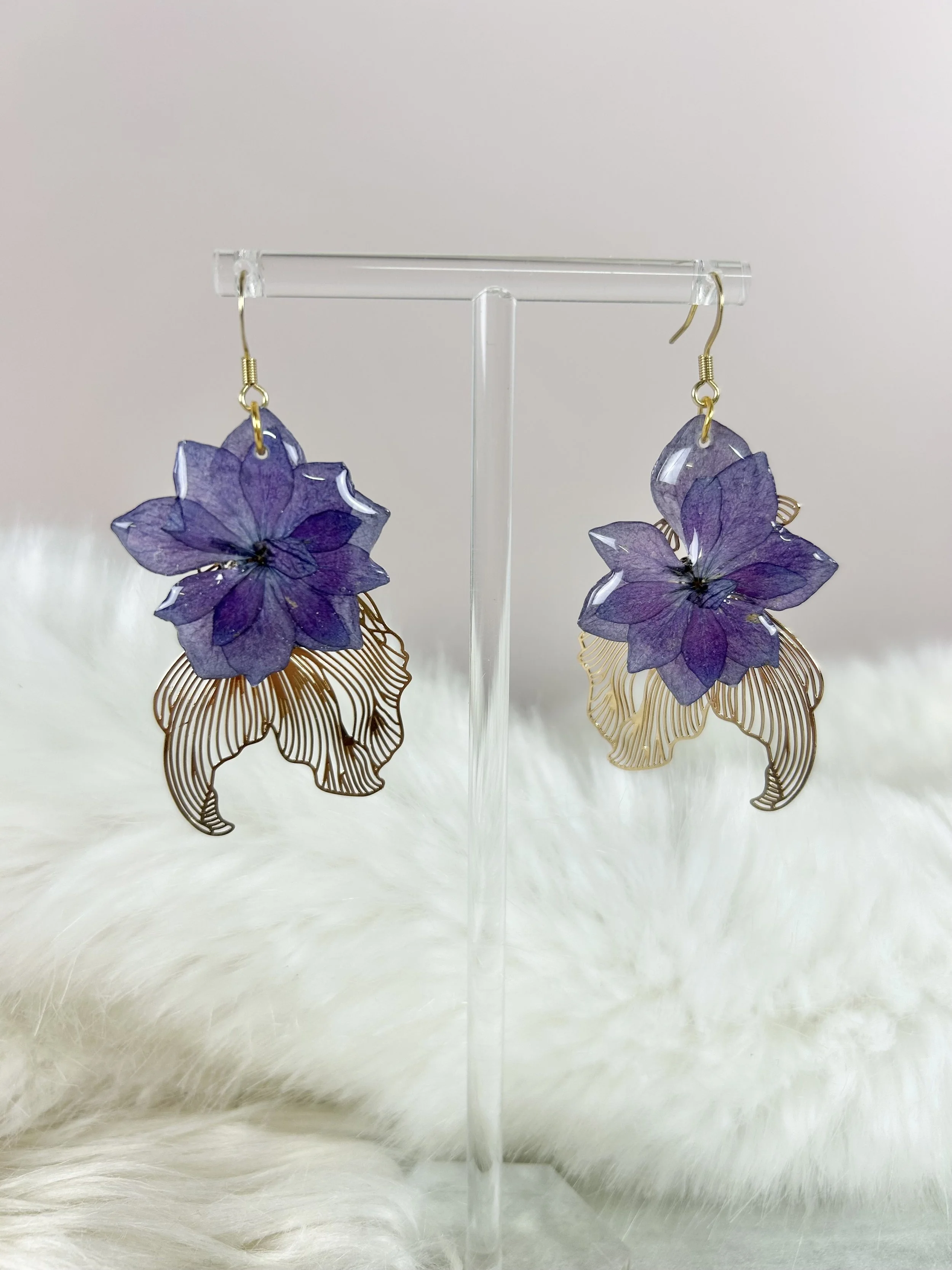 Boucles d'oreilles avec des fleurs violettes en papier ou en tissu, suspendues à des attaches en or, exposées sur un support transparent en forme d'archet.