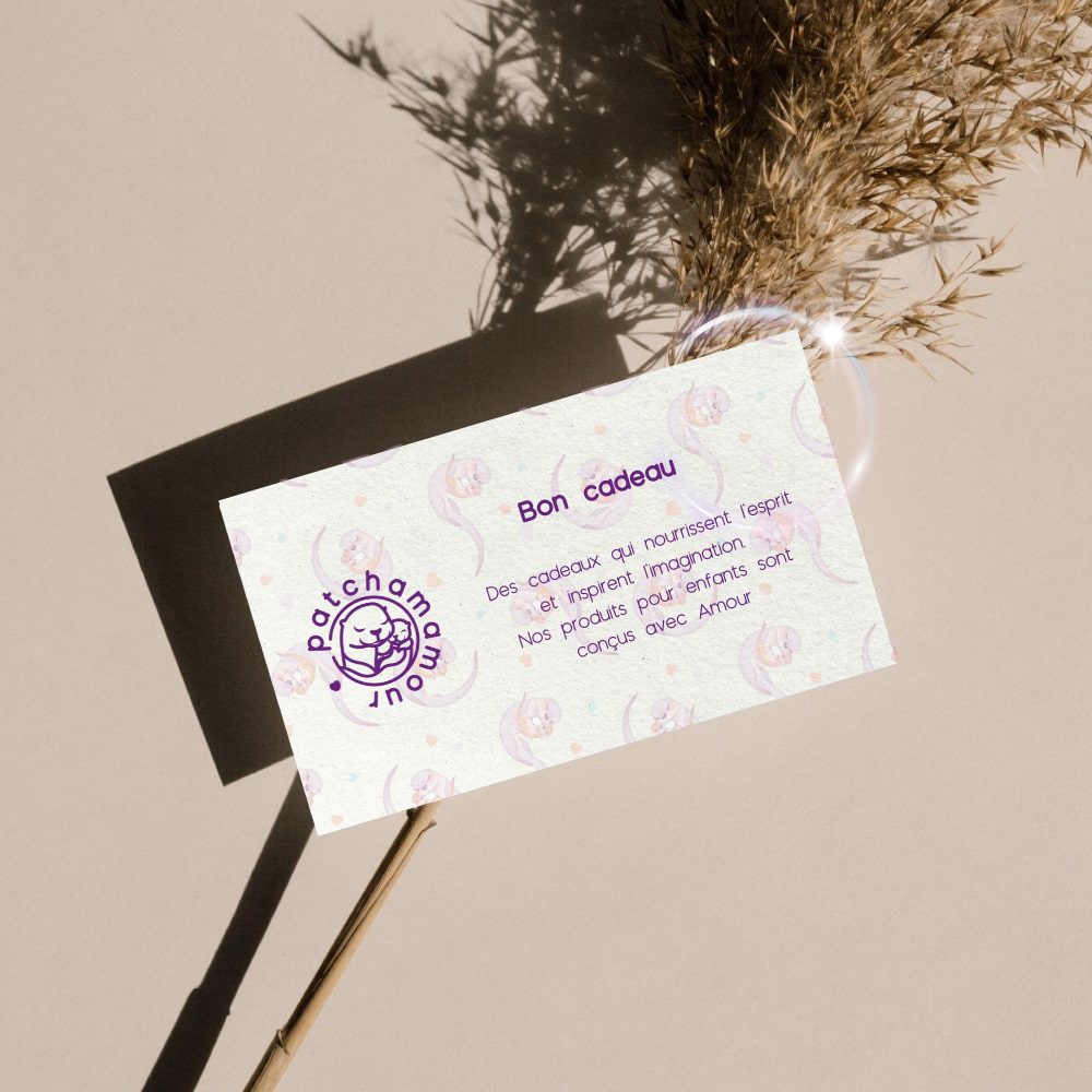 Une carte cadeau avec le texte 'Bon Cadeau' et une description en français sur des produits conçus avec amour, accompagnée d'un logo violet d'une main tenant un cœur. La carte repose sur une branche sèche avec une ombre projetée contre un mur beige, et il y a une petite boule de verre avec des plantes séchées.
