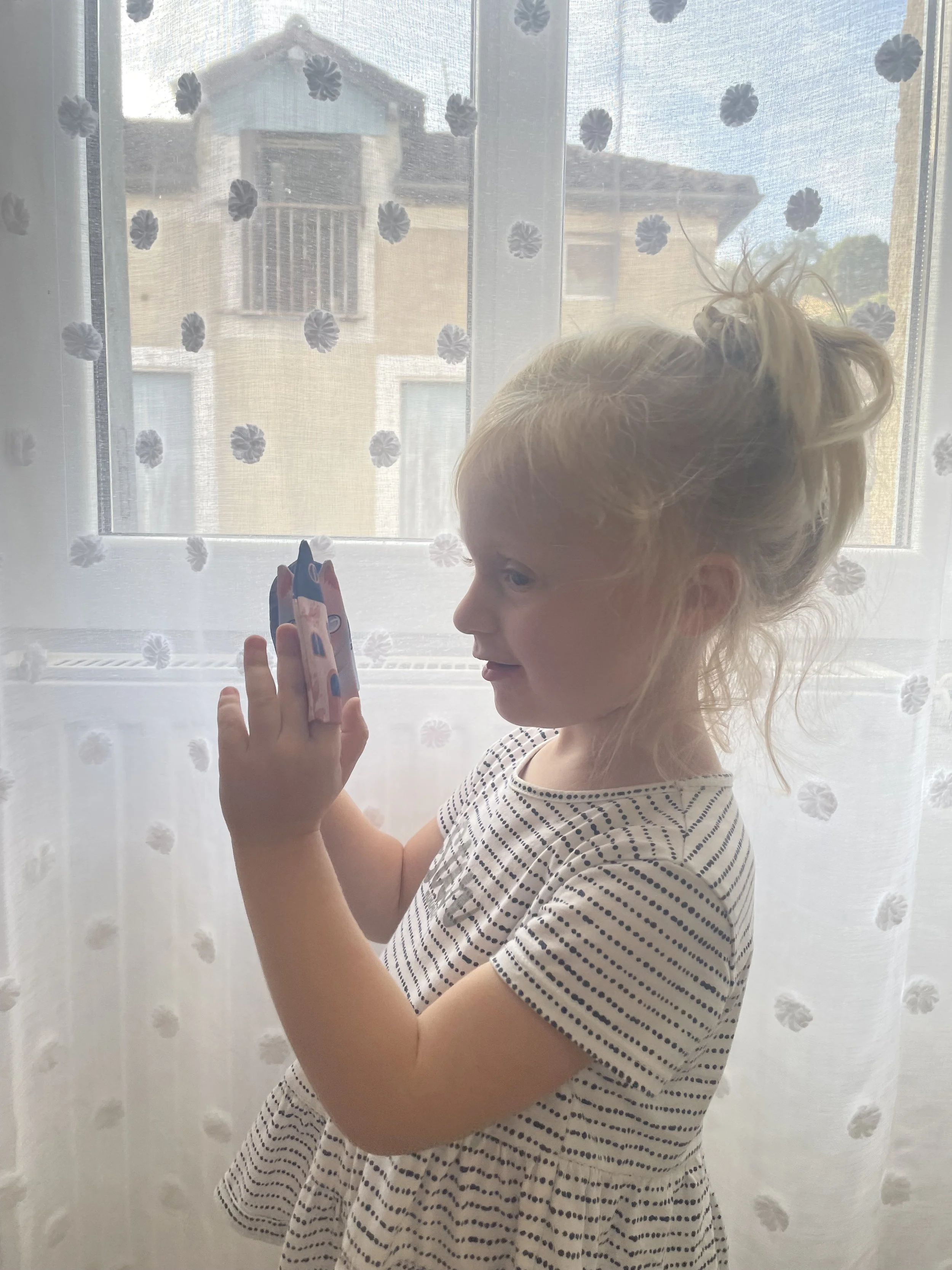 Une petite fille avec les cheveux blonds attachés en chignon regarde un petit objet tenu dans sa main, devant une fenêtre avec des rideaux transparents ornés de motifs floraux, laissant passer la lumière naturelle.