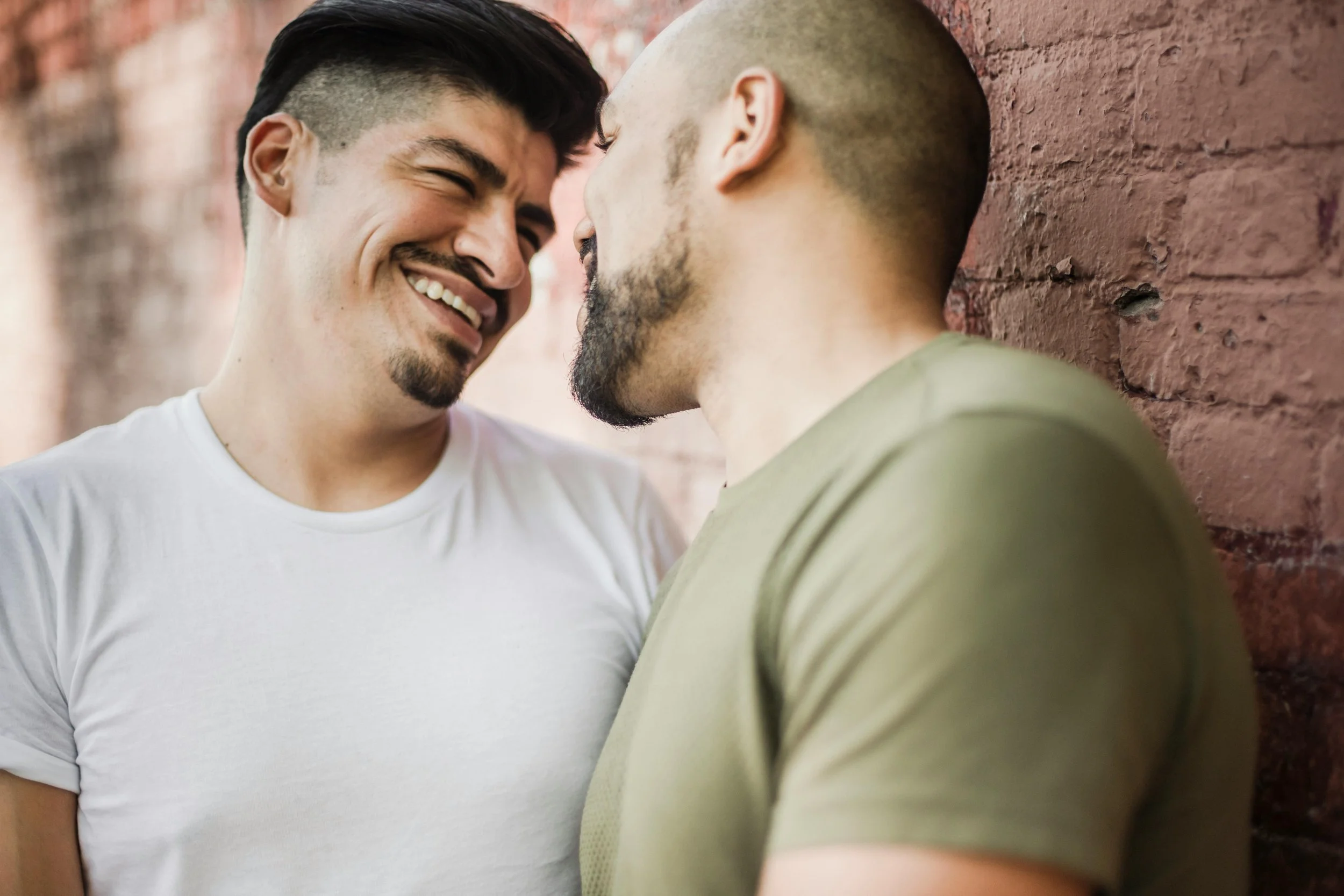 Two men smiling closely, leaning against a brick wall.