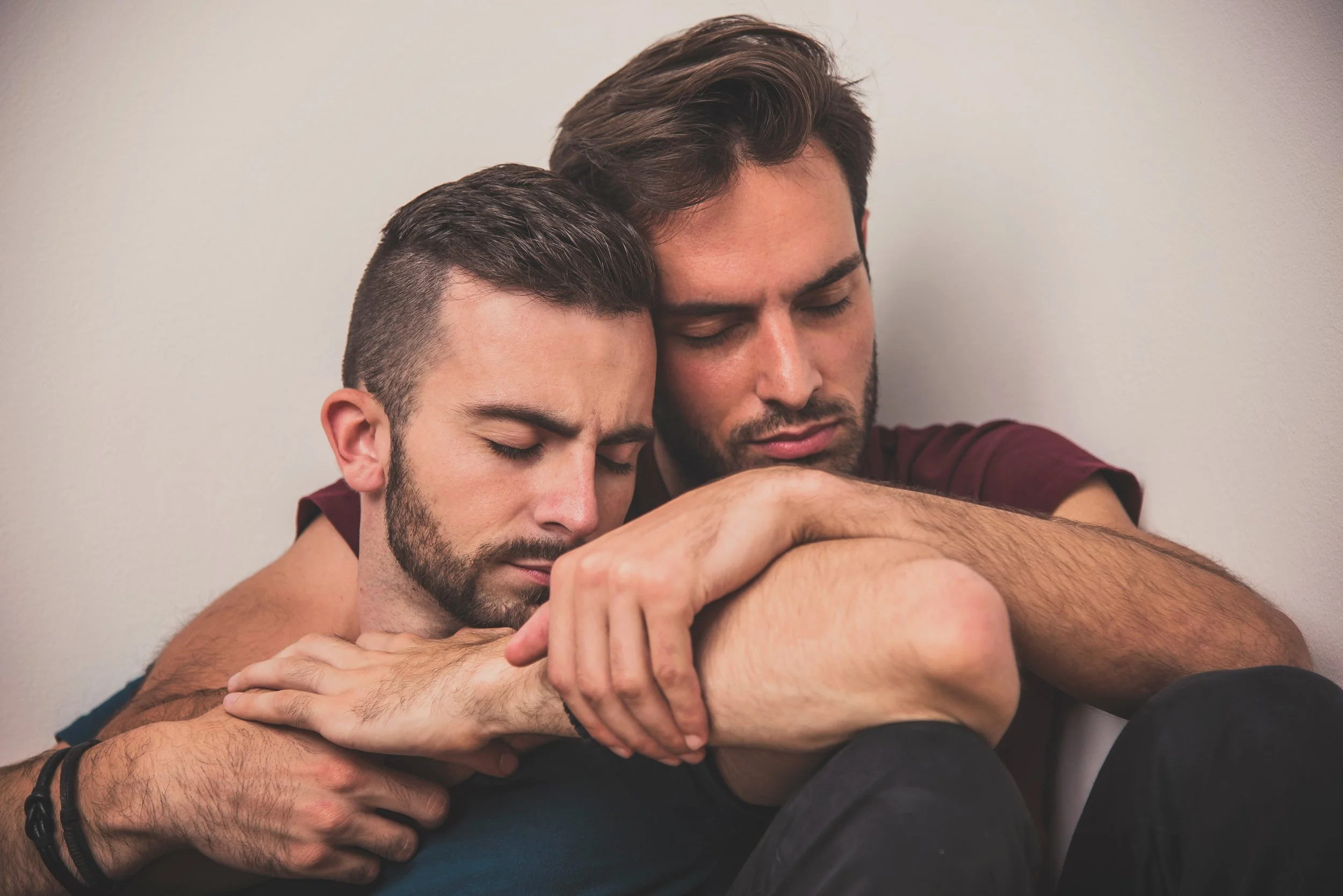 Two men embracing, sitting against a wall, eyes closed, showing affection and comfort.