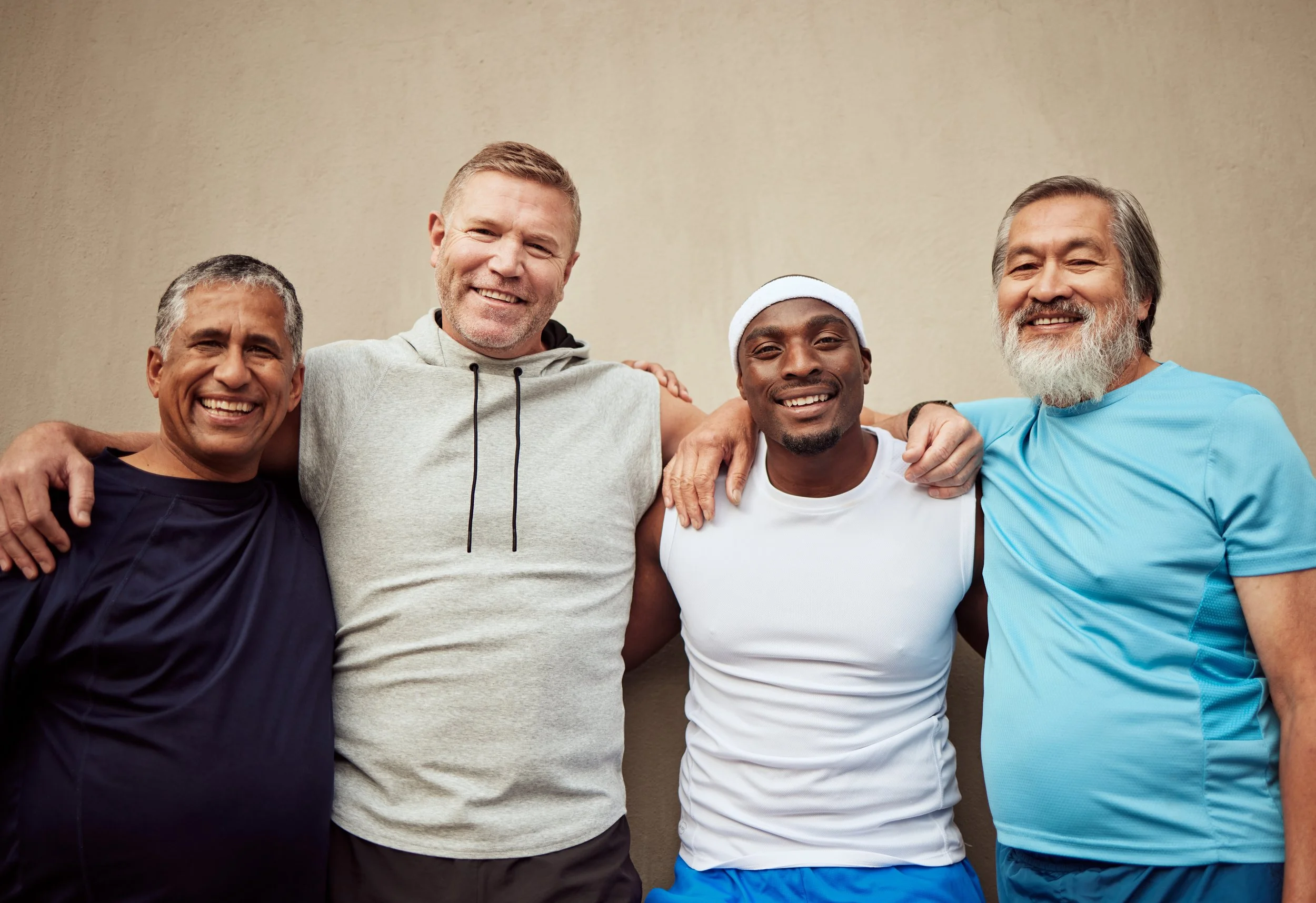 Four smiling men with arms around each other standing against a beige wall.