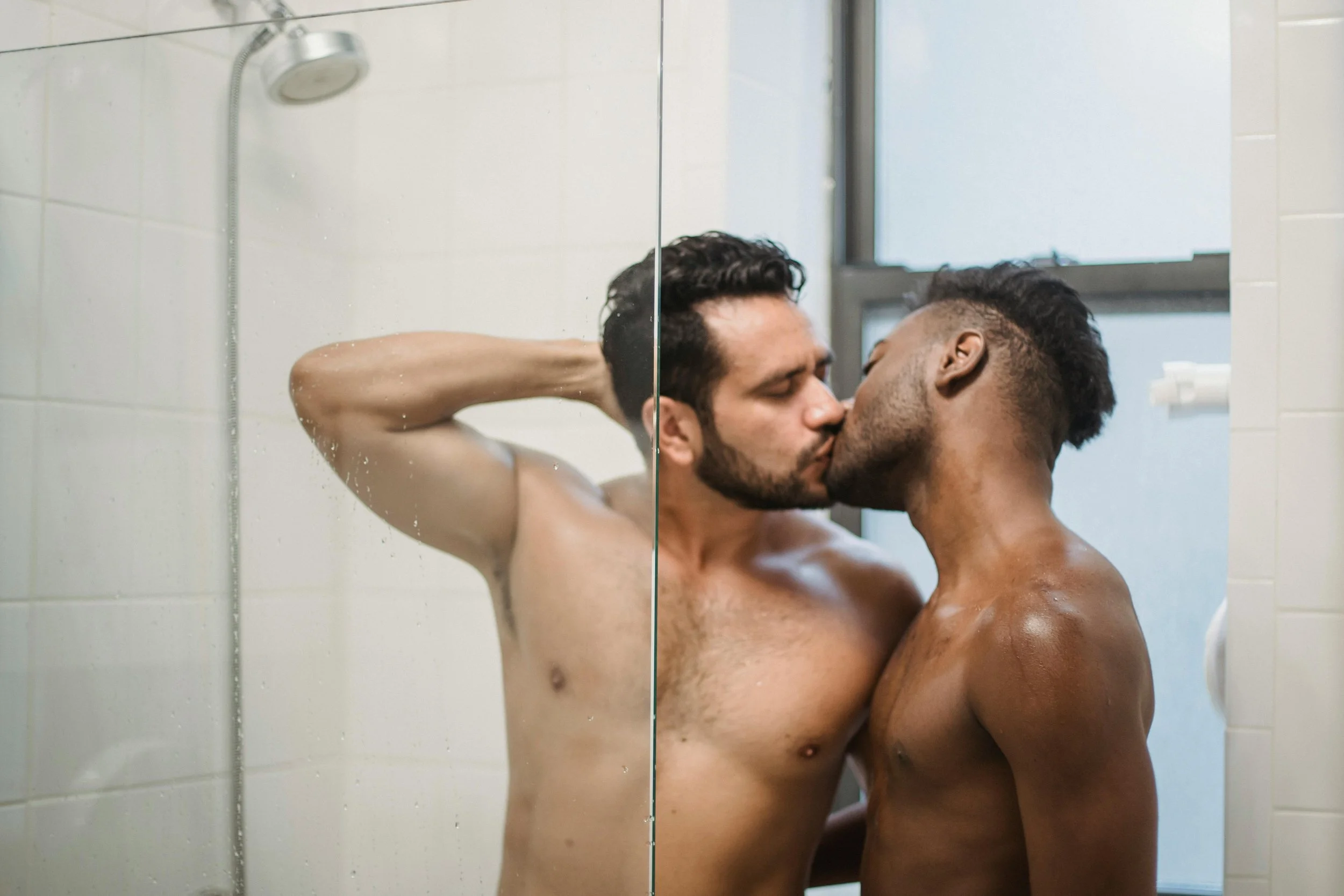Two shirtless men, one with light skin and dark hair and the other with dark skin and a short haircut, sharing a kiss in a bathroom, with a mirror reflecting their intimate moment.