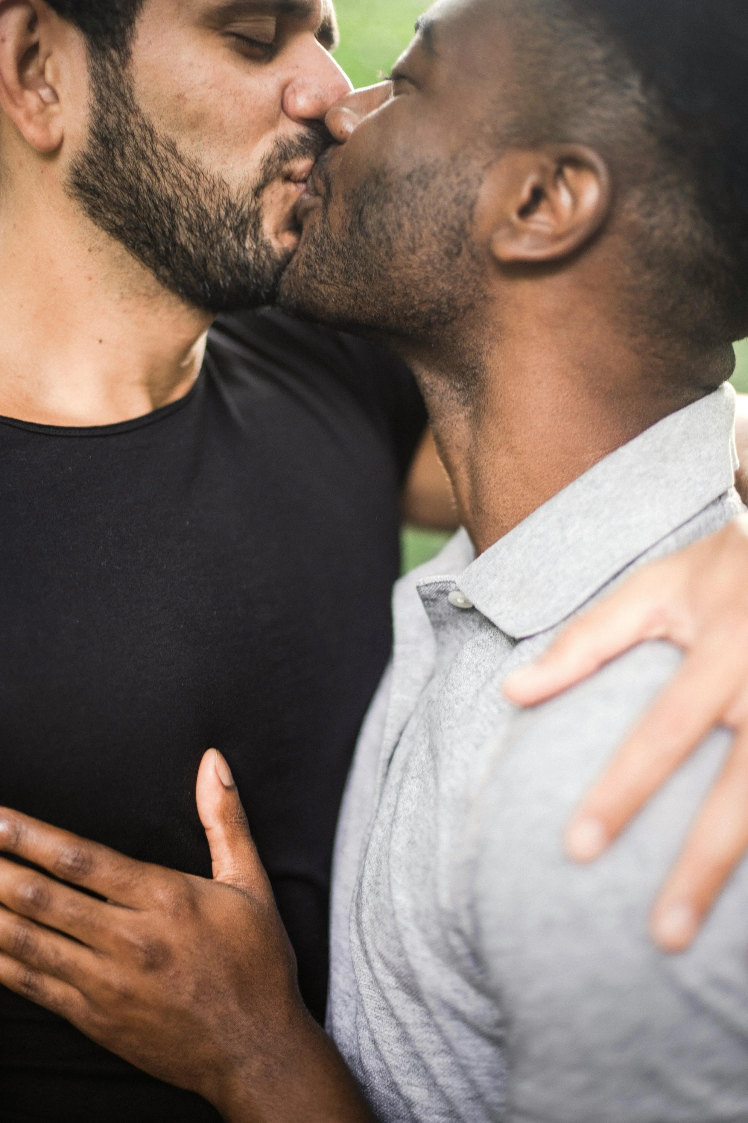 Two men kissing closely, one with a beard and wearing a black shirt, the other with dreadlocks and wearing a gray polo shirt, outdoors.