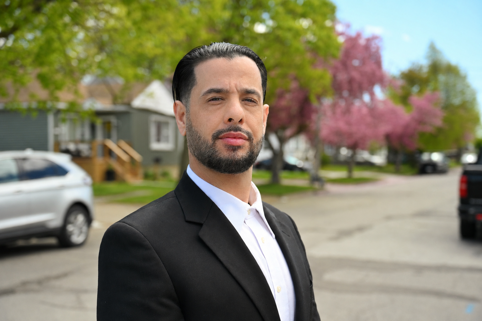 A man with dark hair and beard wearing a black suit and white shirt standing outdoors on a residential street with trees and parked cars, some pink blossoms visible in background.