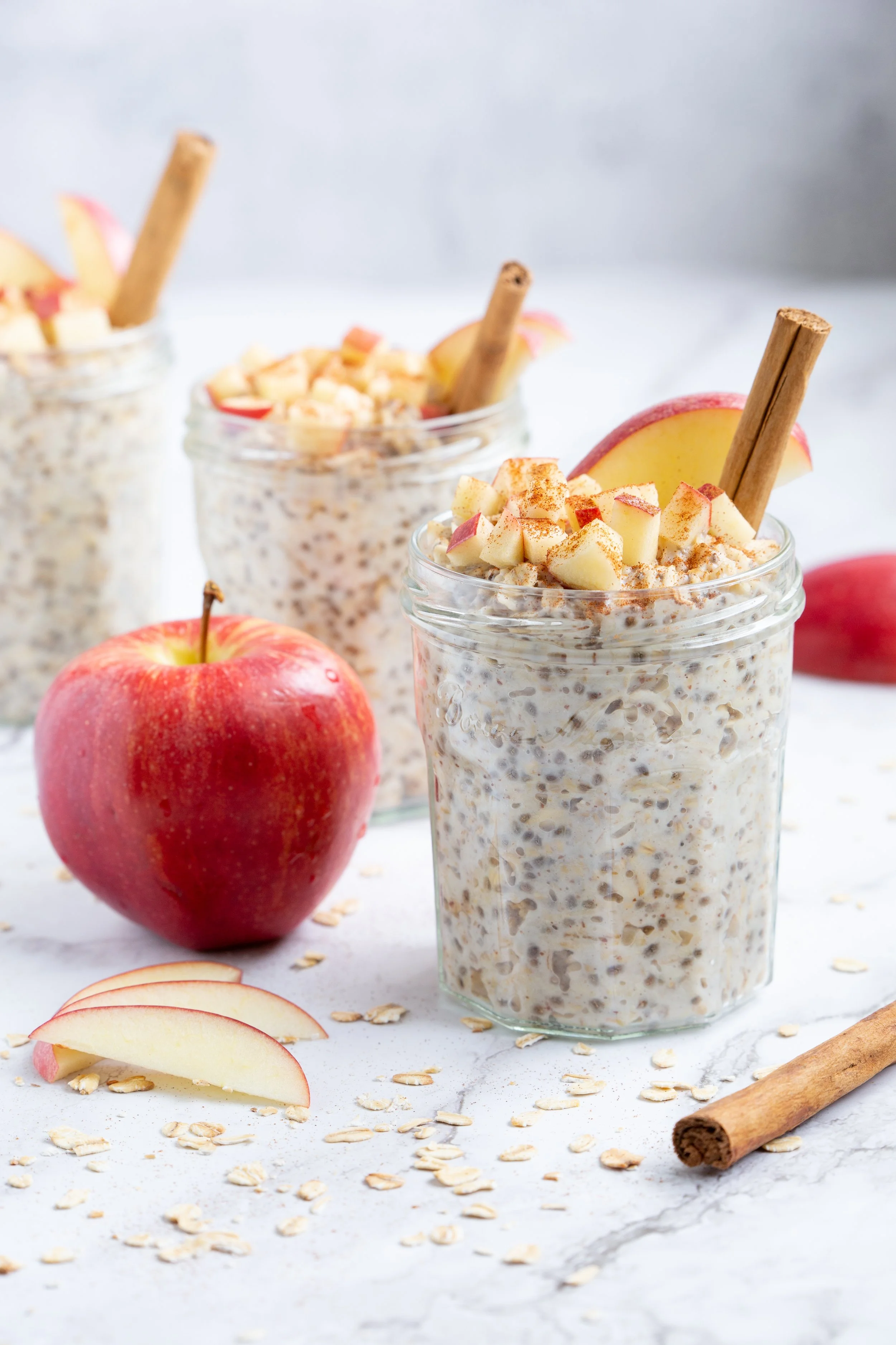 3 jars of overnight oats with diced apples and a cinnamon stick on top. There is a cinnamon stick, apples, and apple slices around the 3 jars.