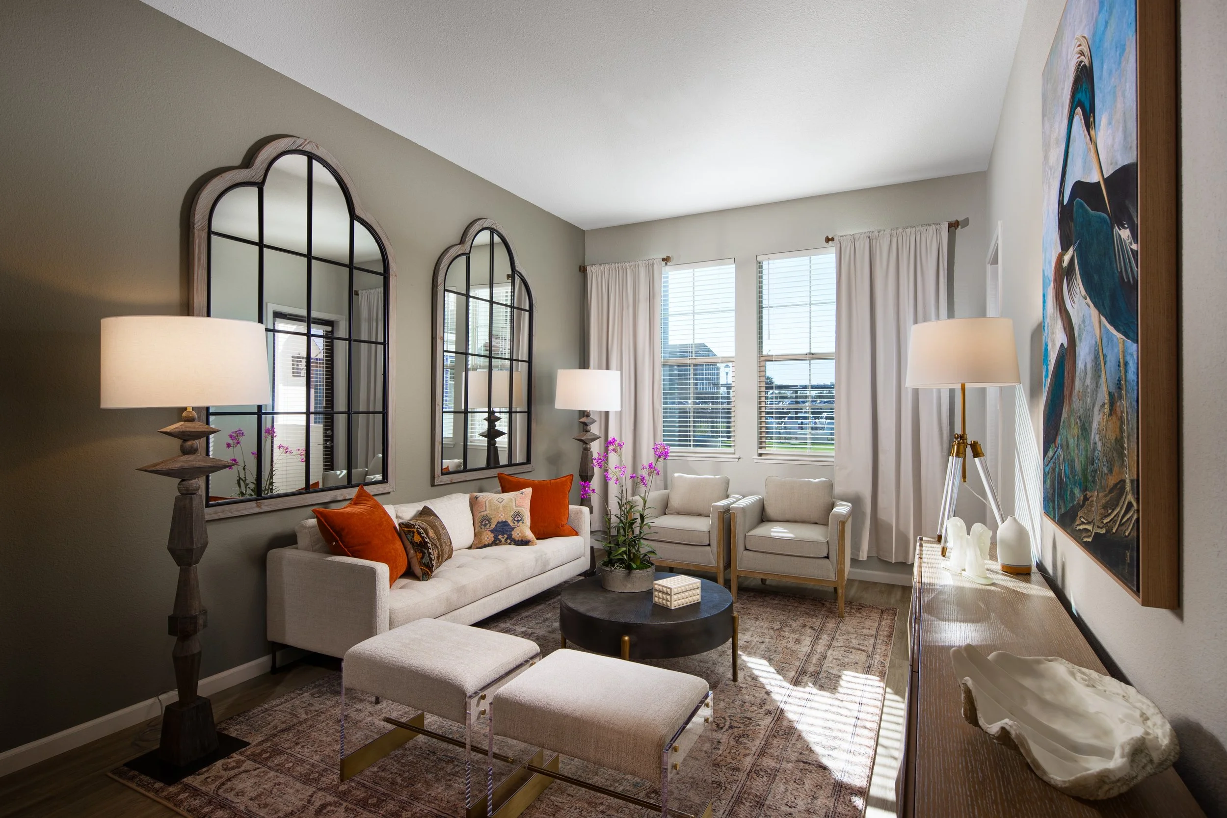 Bright living room with beige sofa, white armchairs, side tables, modern lamps, colorful pillows, large windows with white blinds and curtains, artwork of a bird on the wall, decorative mirror panels, and a coffee table with purple orchids.