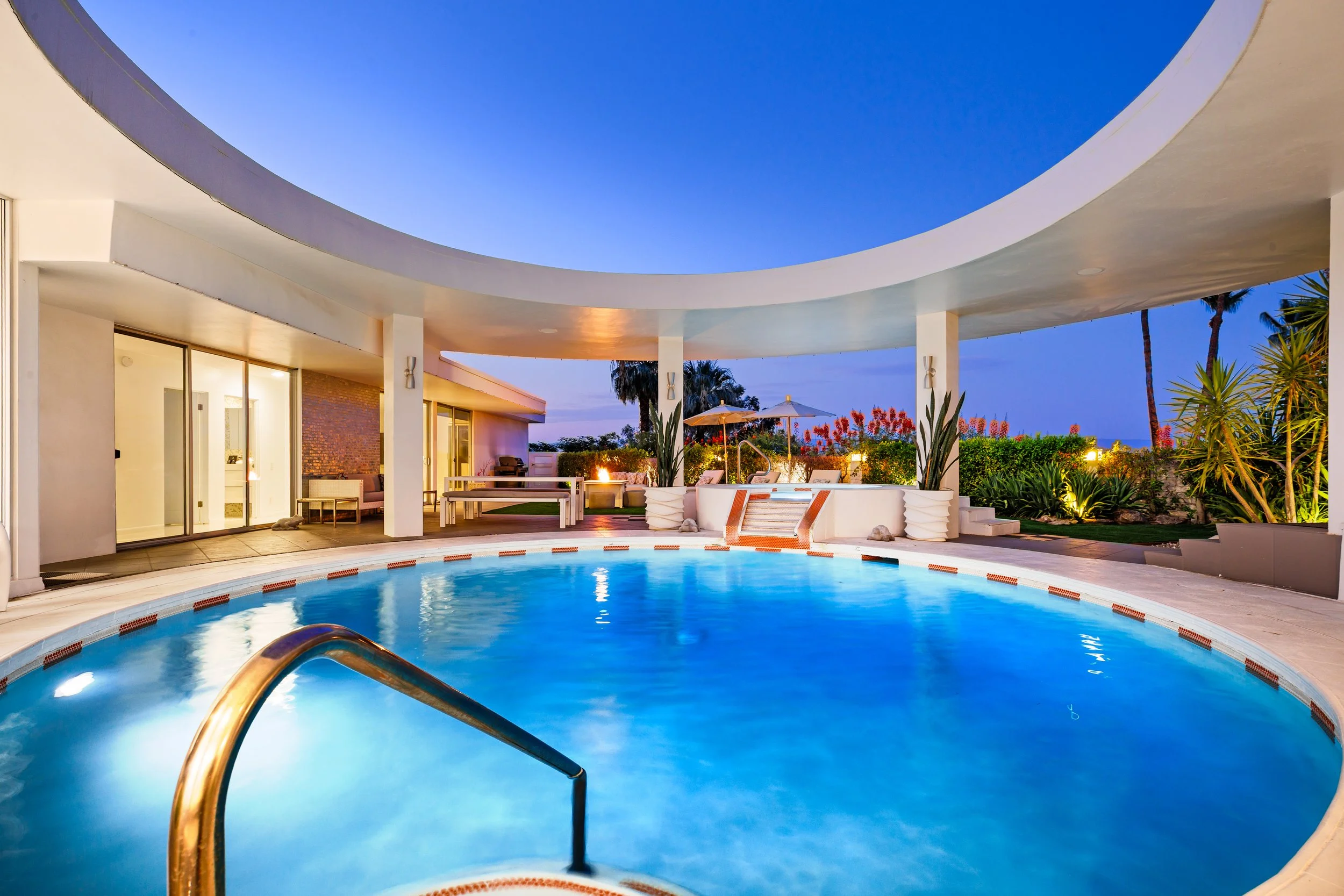 Nighttime view of a modern house with a swimming pool and outdoor seating area, surrounded by plants and palm trees.