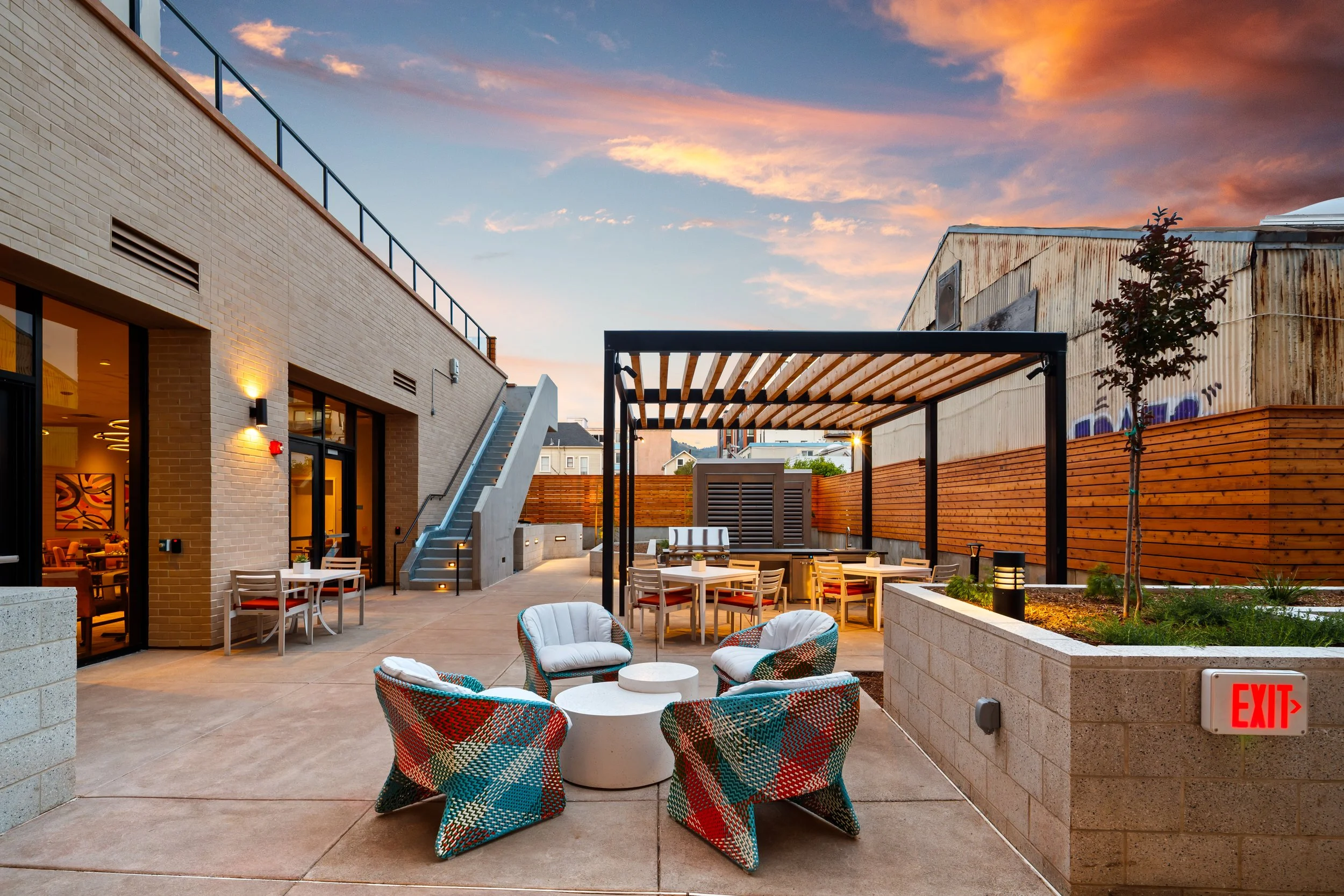 Outdoor patio area at sunset with chairs, tables, and a pergola, surrounded by a brick building on the left and a wooden fence on the right.