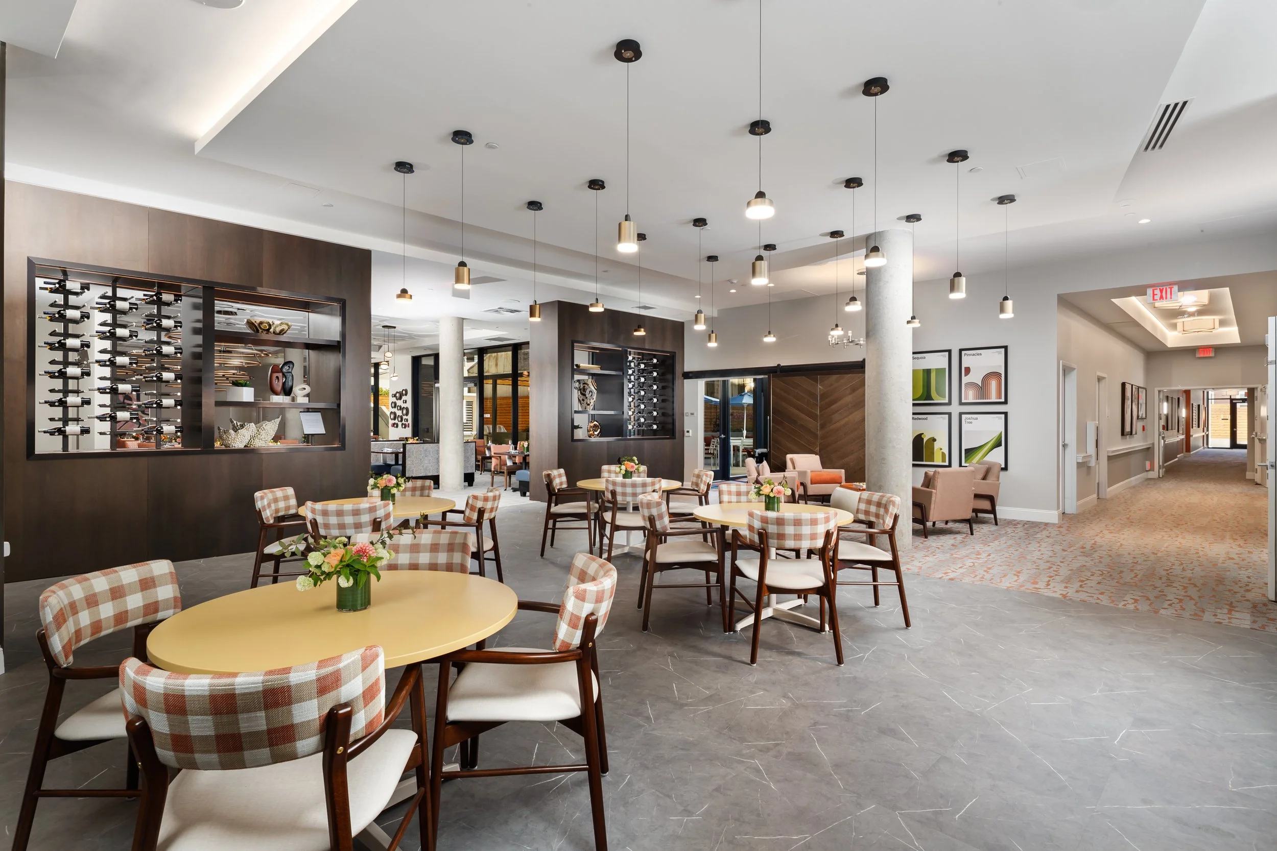 A spacious restaurant or lounge area with round tables, floral centerpieces, checkered chairs, modern lighting, wine display, artwork on walls, and carpeted hallways.
