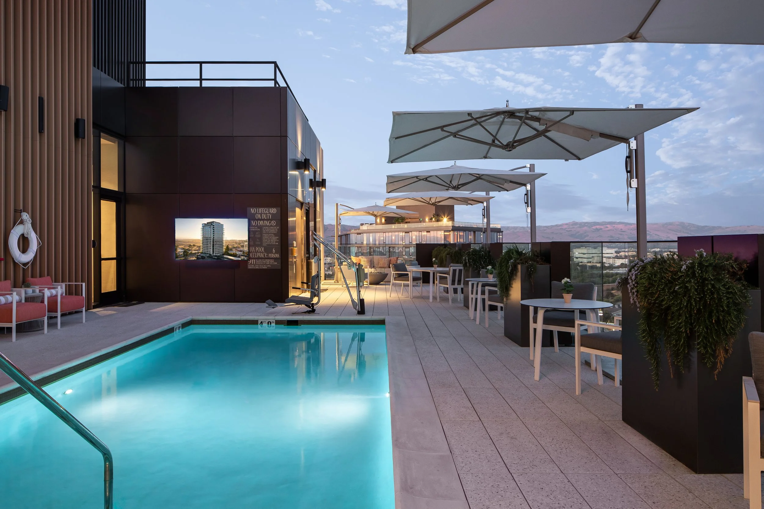 Rooftop swimming pool area with tables, chairs, umbrellas, and potted plants, overlooking a cityscape at dusk.