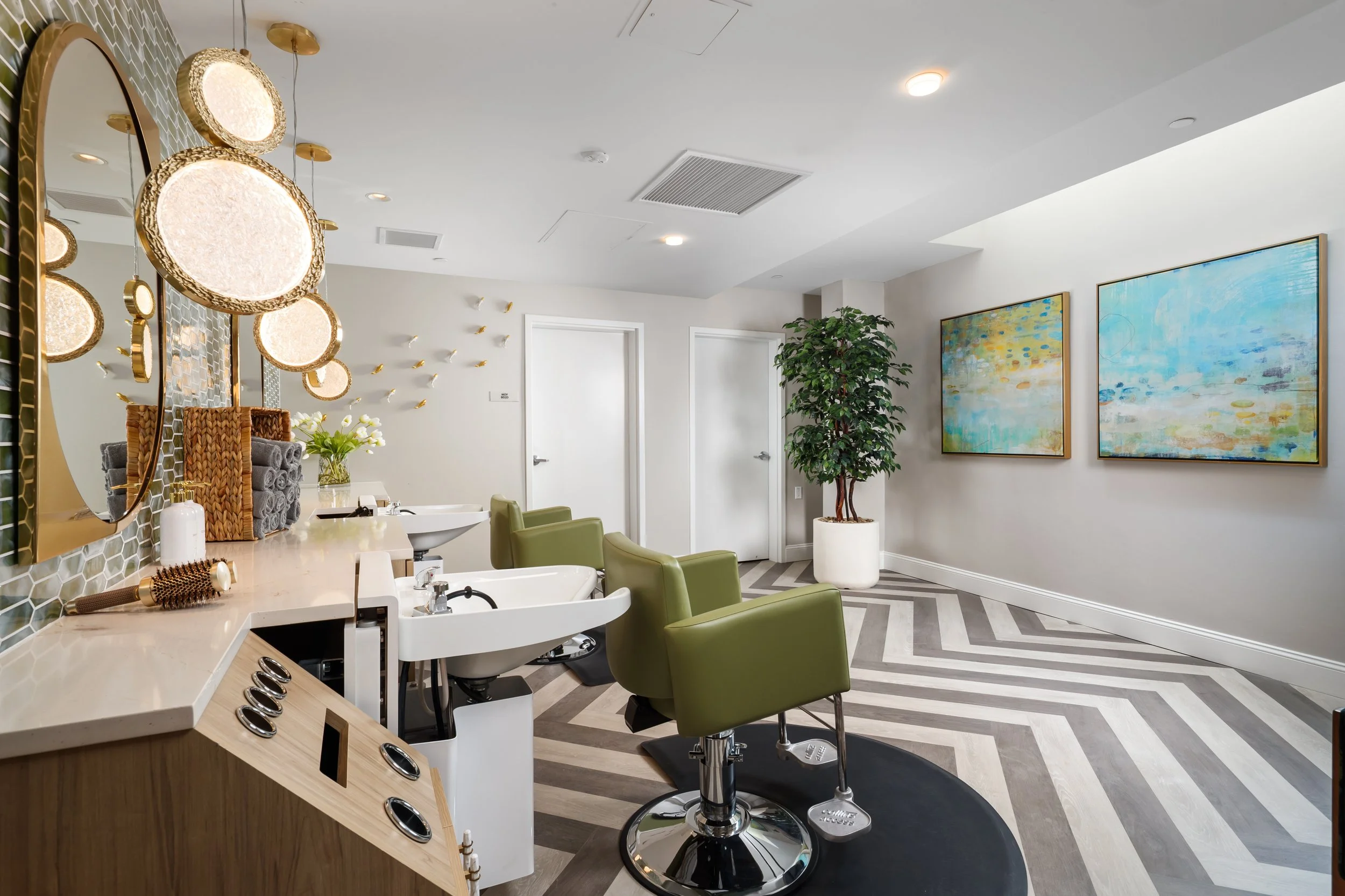 Interior view of a modern hair salon with green chairs, white washbasins, wall art, large mirror, and decorative lighting, featuring a chevron patterned floor and a tall potted plant.