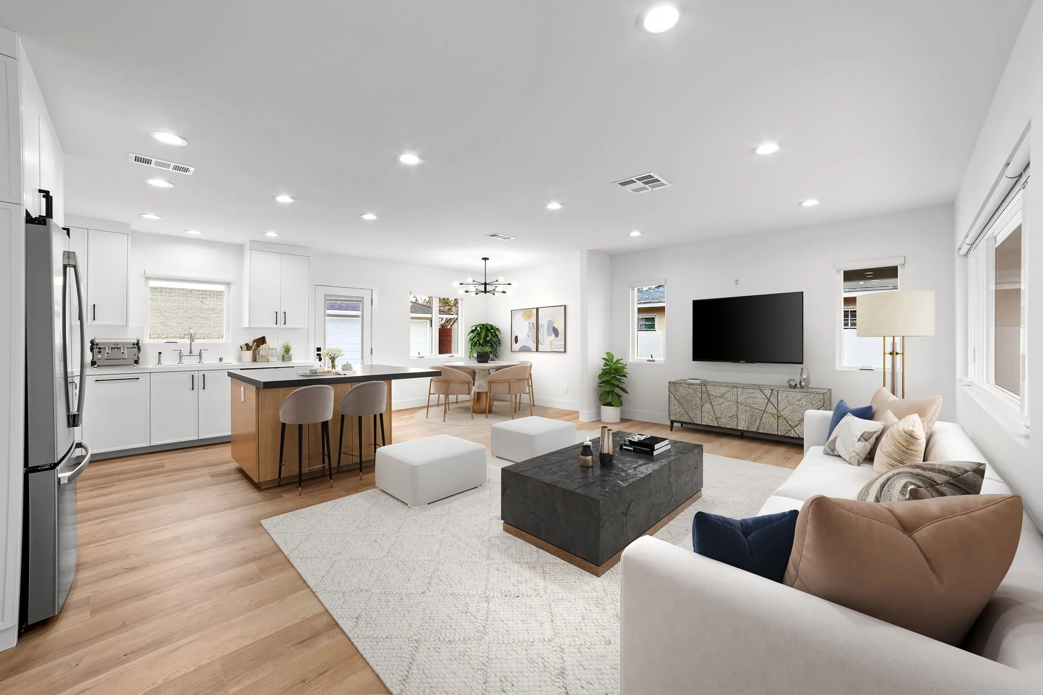 Open-concept living room with white walls and ceiling, hardwood floors, a large flat-screen TV, beige and white sofas, a black coffee table, a dining table with pink chairs, kitchen with white cabinets, window with blinds, and modern lighting fixtures.
