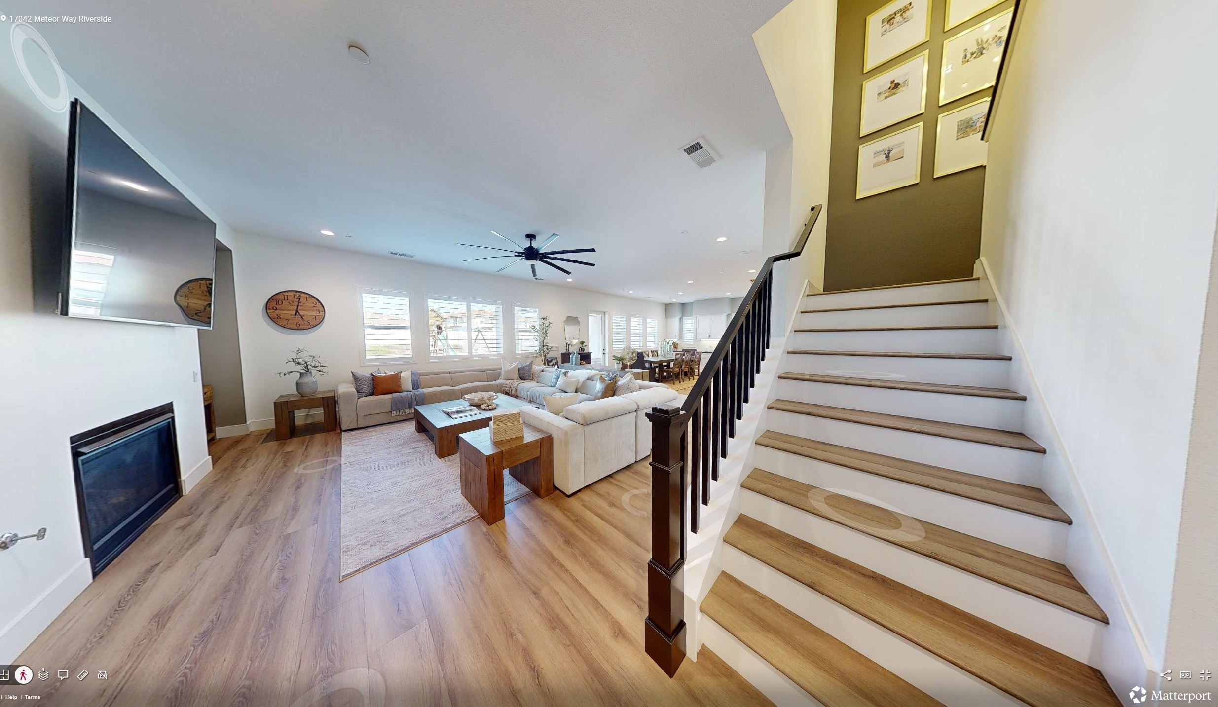 Living room with beige L-shaped sofa, wooden coffee table, TV mounted on white wall, black ceiling fan, multiple windows with white blinds, framed pictures on the green accent wall, staircase with wooden steps and black railing, hardwood flooring, and dining area in the background.