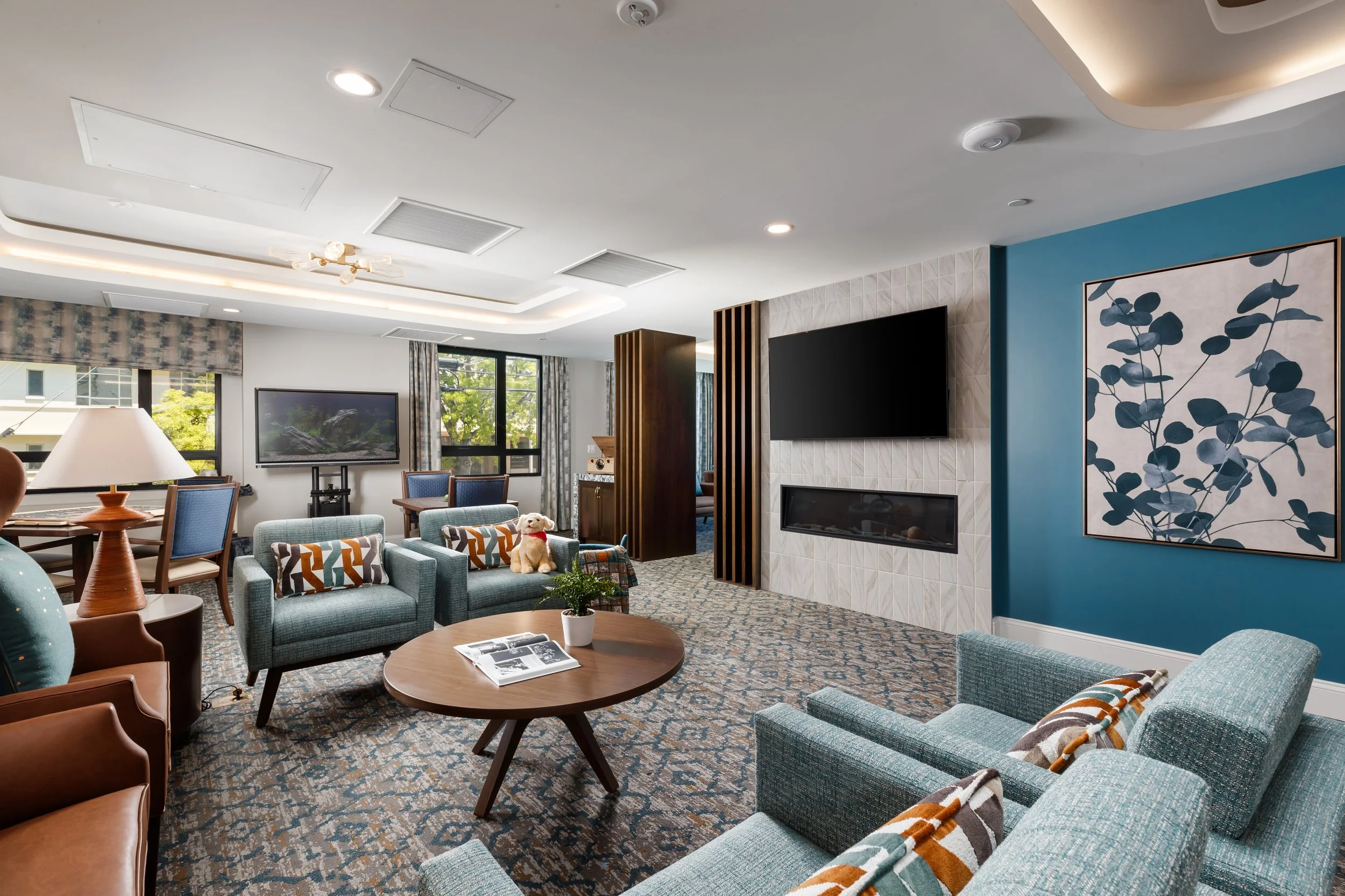 Modern living room with blue walls, patterned carpet, and furniture including two armchairs, a sofa, a round coffee table with magazines and a plant, and a television mounted on a tiled wall with a fireplace below. Large windows with curtains, and de