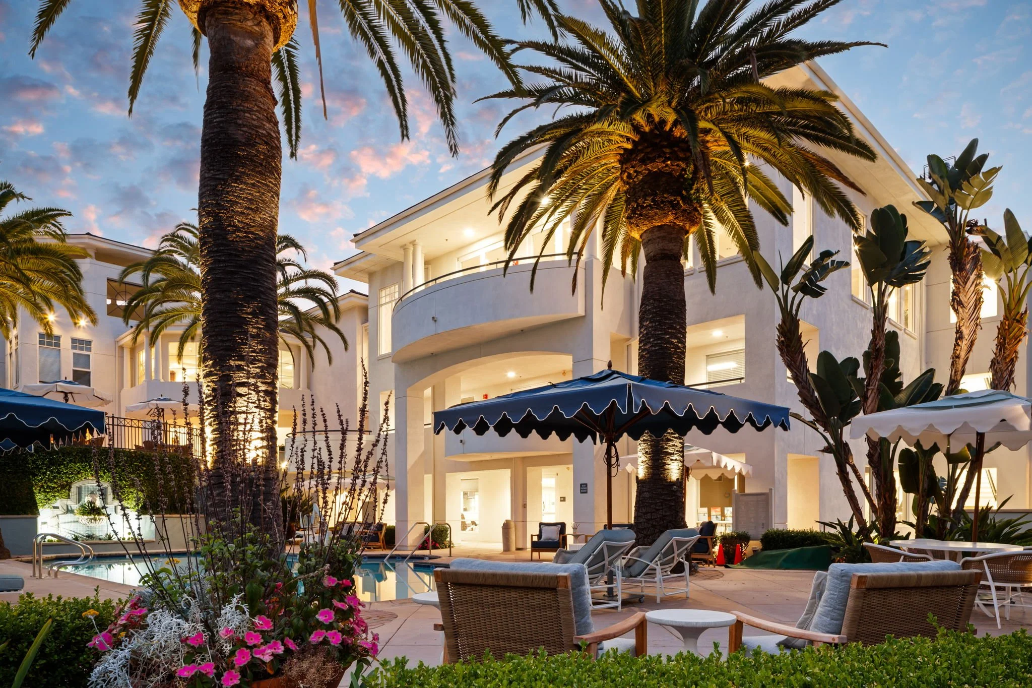 A luxury resort with white multi-story buildings, palm trees, outdoor seating, umbrellas, a pool, and colorful flowers, during early evening with sky transitioning to night.