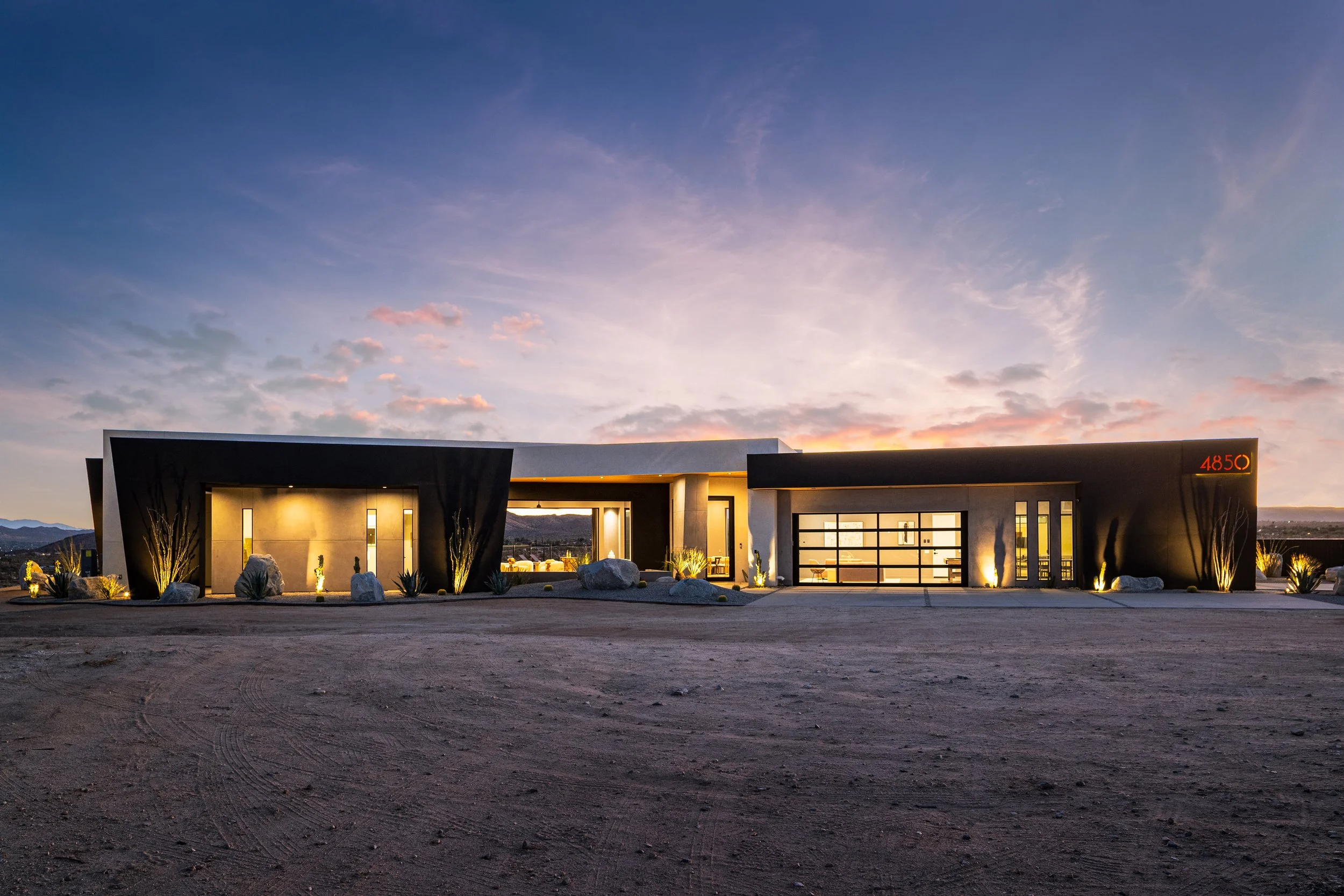 Modern house at dusk with large glass windows, desert landscaping, rocks, and minimalistic lighting.