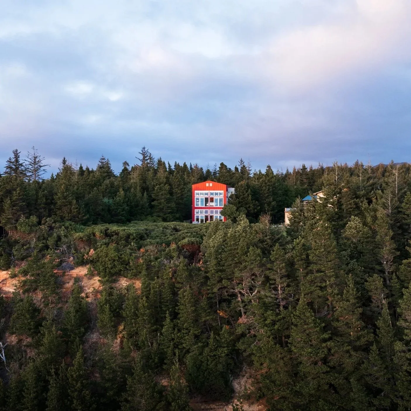 A red house on a hill surrounded by dense trees under a cloudy sky.