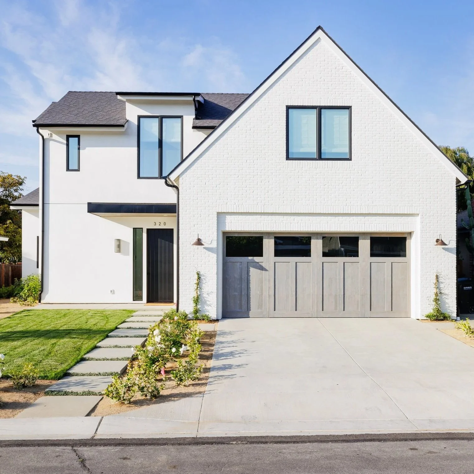 Modern two-story white house with gray roof, attached garage, front lawn, and garden with flowers and shrubs under a blue sky.