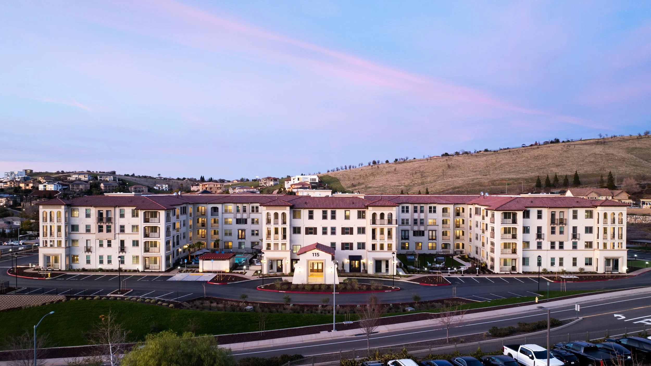 Elevated view of a modern apartment complex with white walls and red-tiled roofs, parking lot in foreground, rolling hills in background during dusk