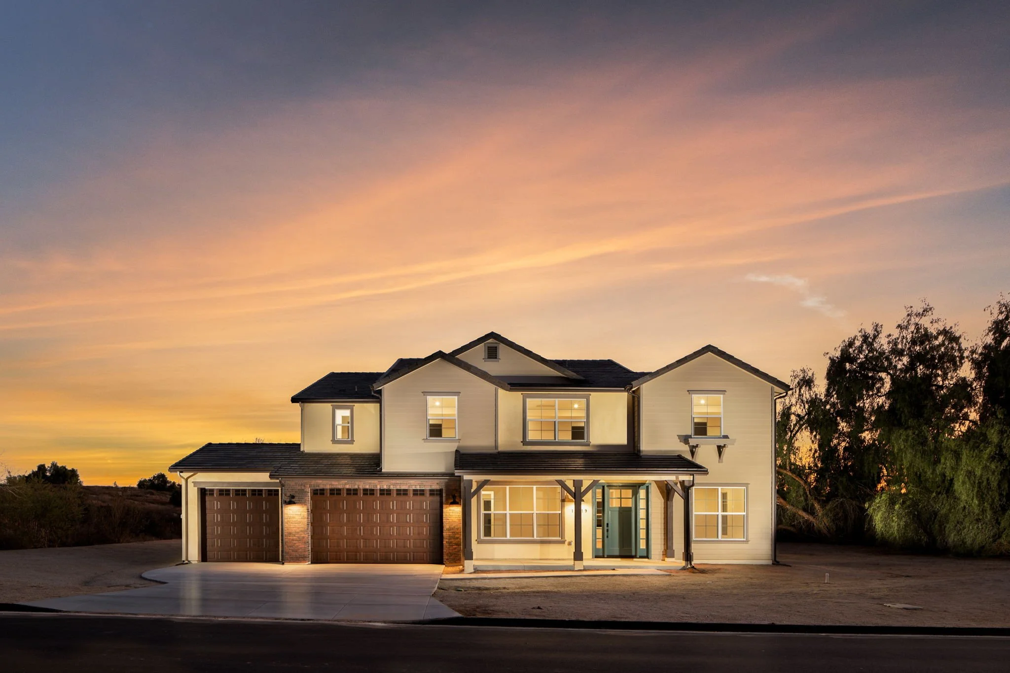 A two-story house with a garage, illuminated from inside, under a colorful sunset sky, with trees around the house.