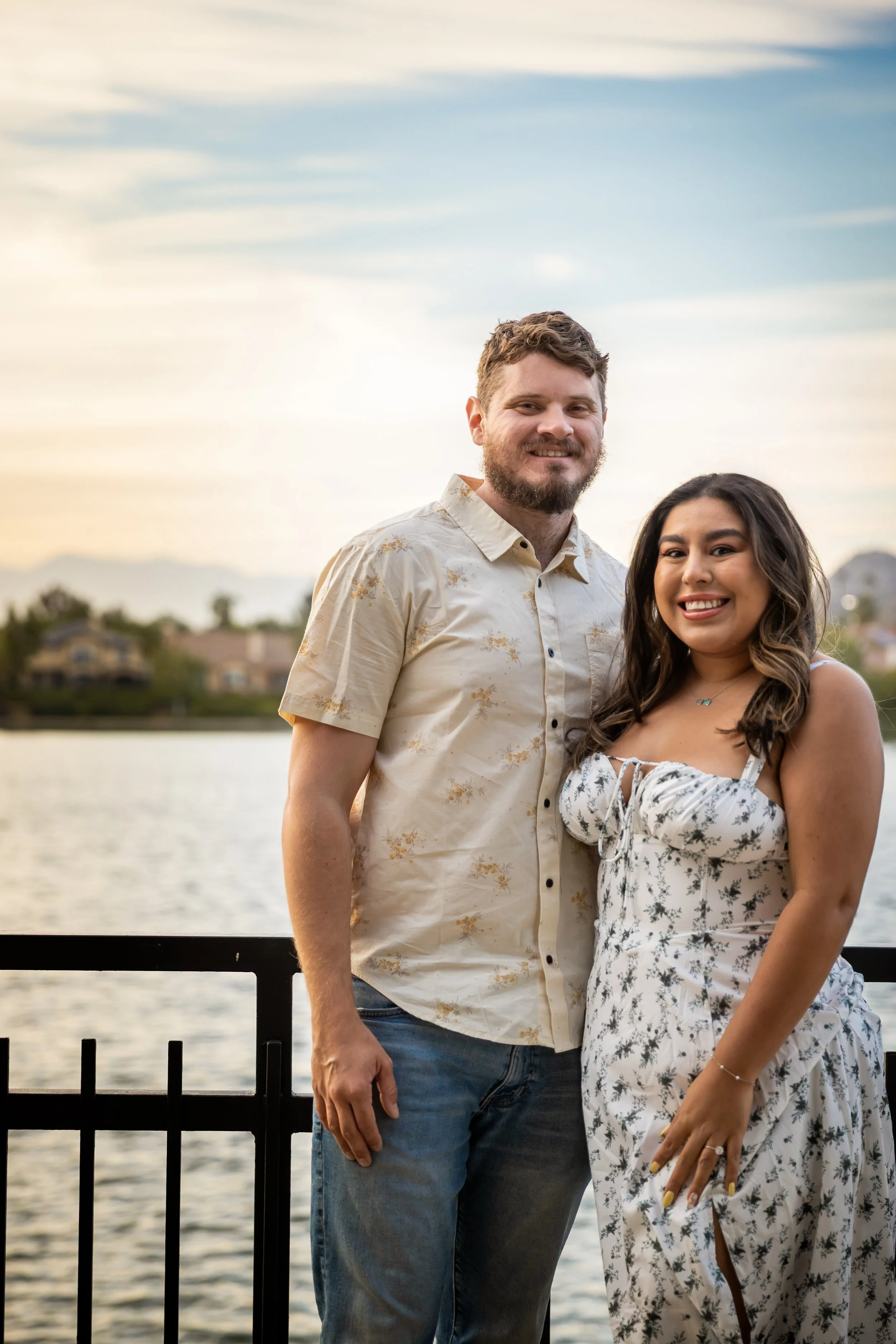 A smiling man and woman standing outdoors near a body of water during sunset, with a cityscape in the background.