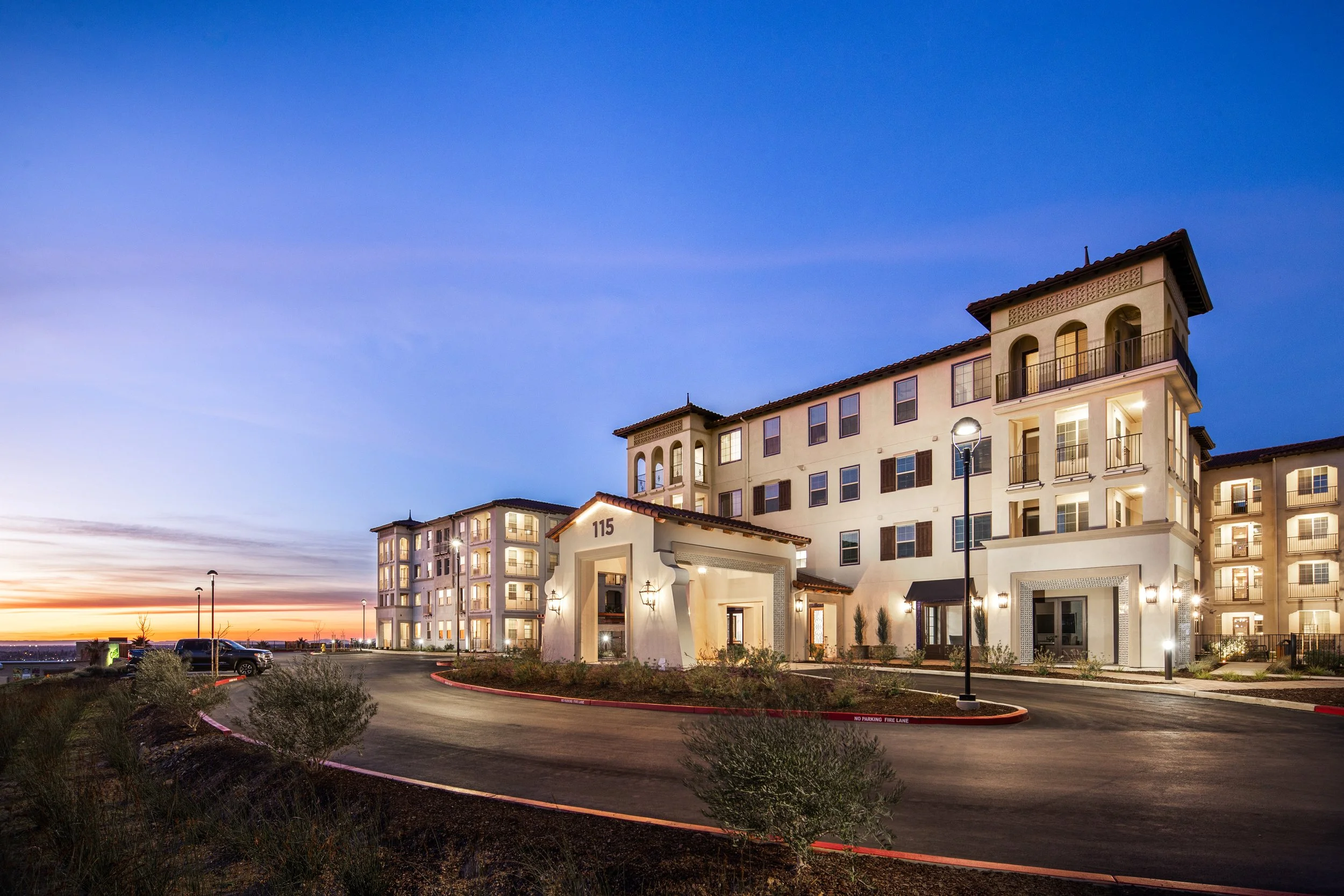 A multi-story apartment complex at sunset with lit exterior lights, a curved driveway, landscaping, and a clear evening sky.