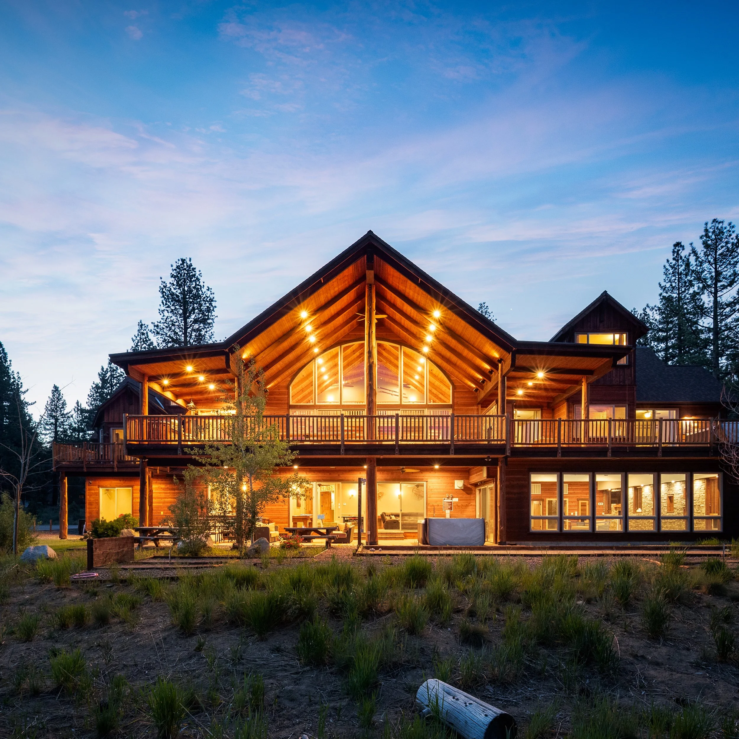 Large two-story wooden house with illuminated windows and a spacious balcony, surrounded by trees and a grassy yard during dusk.