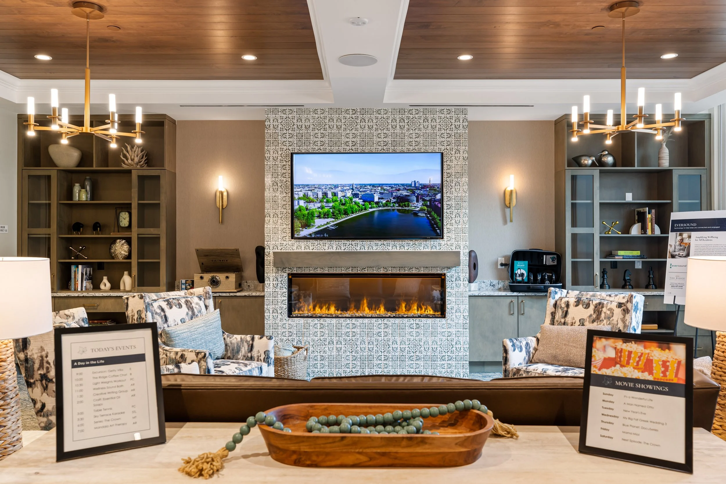 A cozy living room with a modern fireplace, a wall-mounted flat-screen TV showing a cityscape, and stylish seating. The room has warm lighting, built-in shelves, and decorative objects.