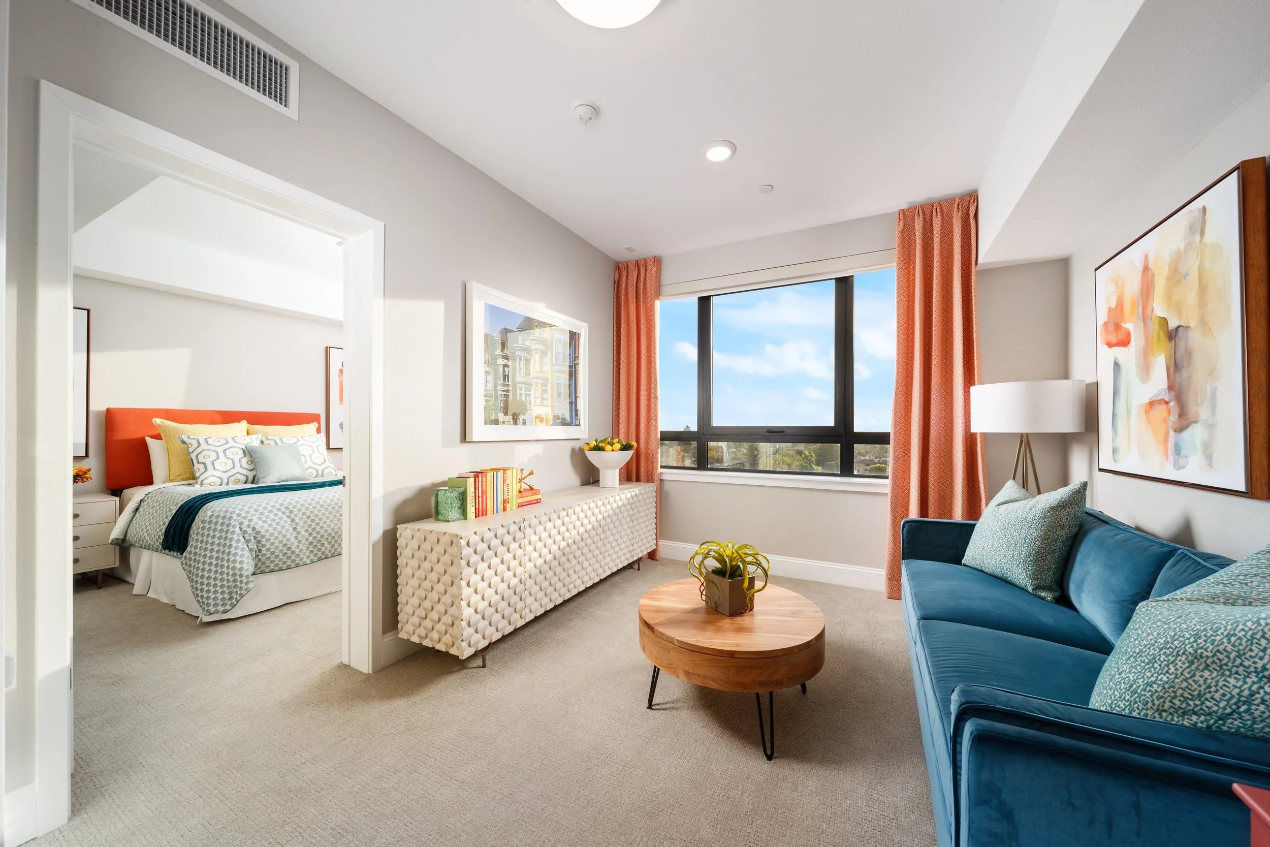 Living room with a blue sofa, a wooden coffee table, and a lamp. Adjacent bedroom with a bed, pillows, and window with orange curtains, depicting a modern apartment.