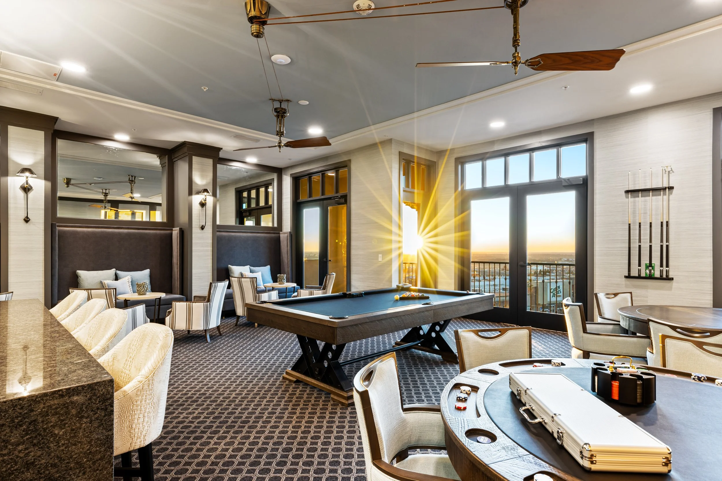 A spacious lounge room with large windows and a sunset view. The room has a pool table, a round poker or card table with poker chips, a case on the table, and comfortable chairs and sofas. Ceiling fans and modern decor are visible.