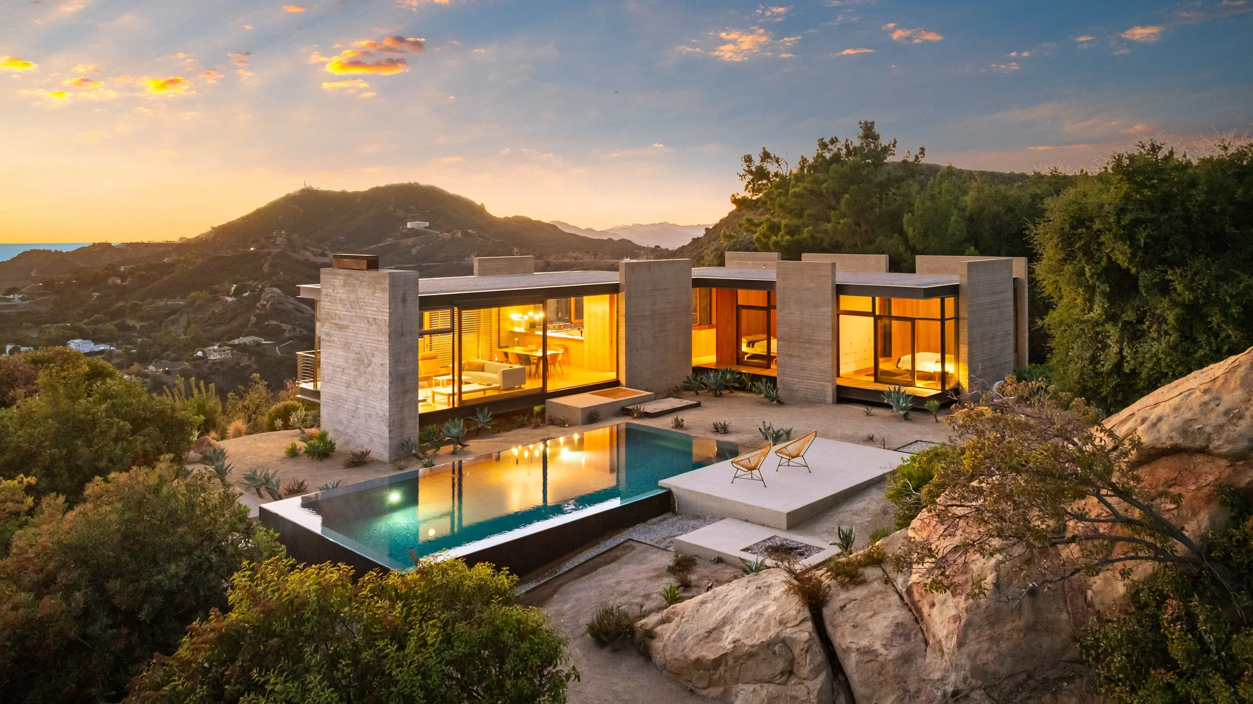 Modern house on hillside with large glass windows, private pool, two outdoor chairs, surrounded by desert plants and rocks, overlooking mountains during sunset.