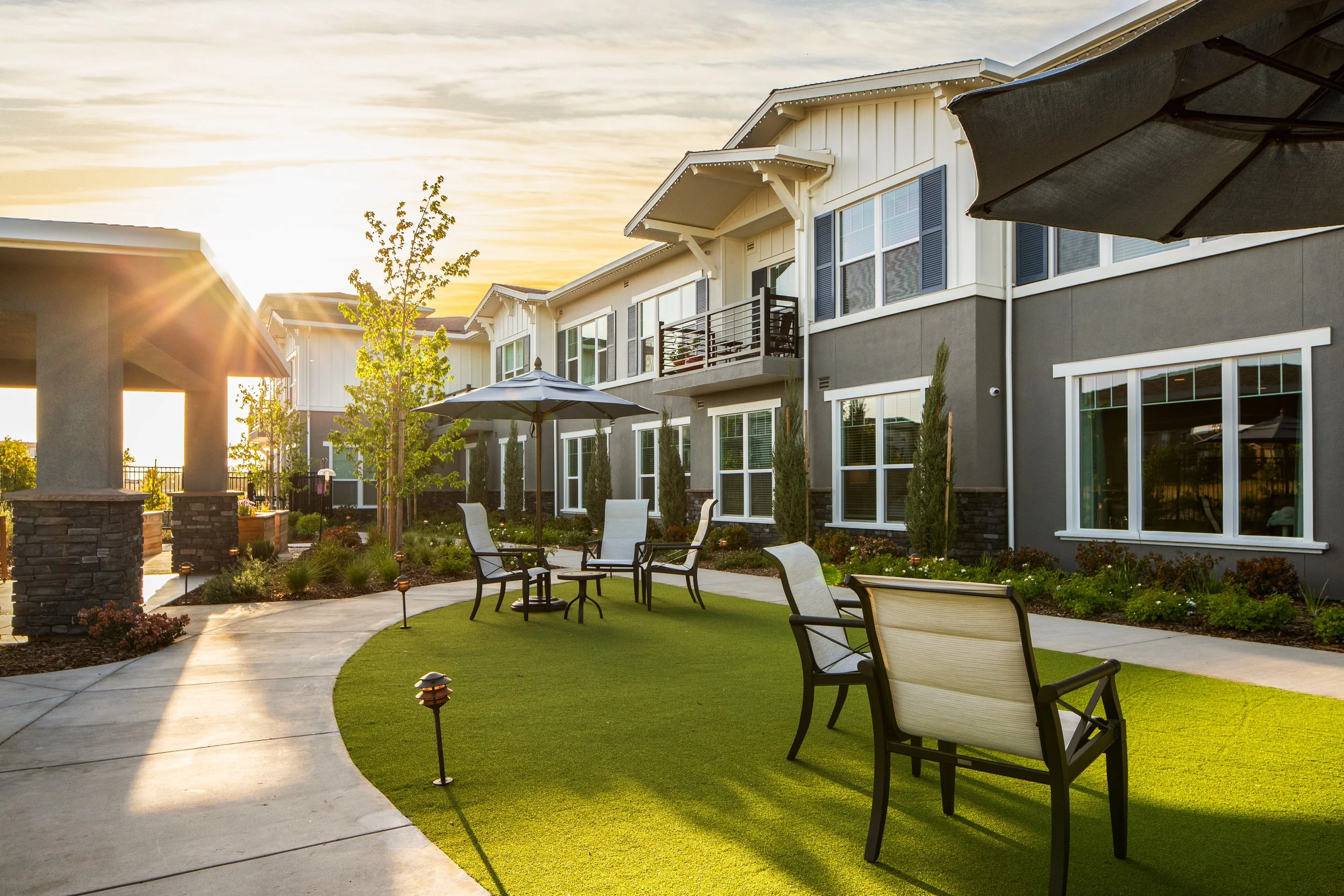 Apartment complex outdoor courtyard at sunset with patio furniture, umbrellas, and landscaped garden.