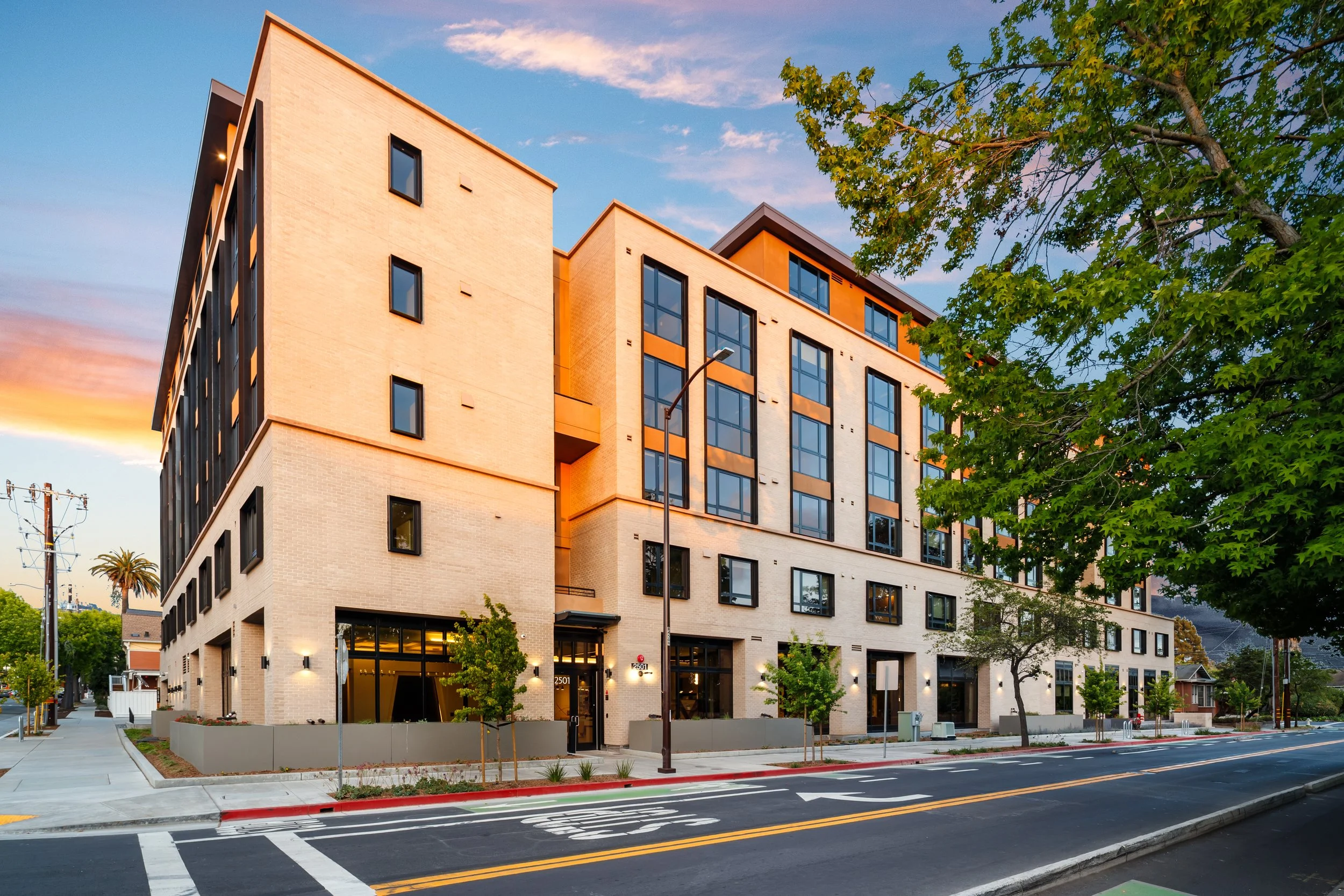 Modern multi-story residential apartment building at sunset with trees and street in foreground.