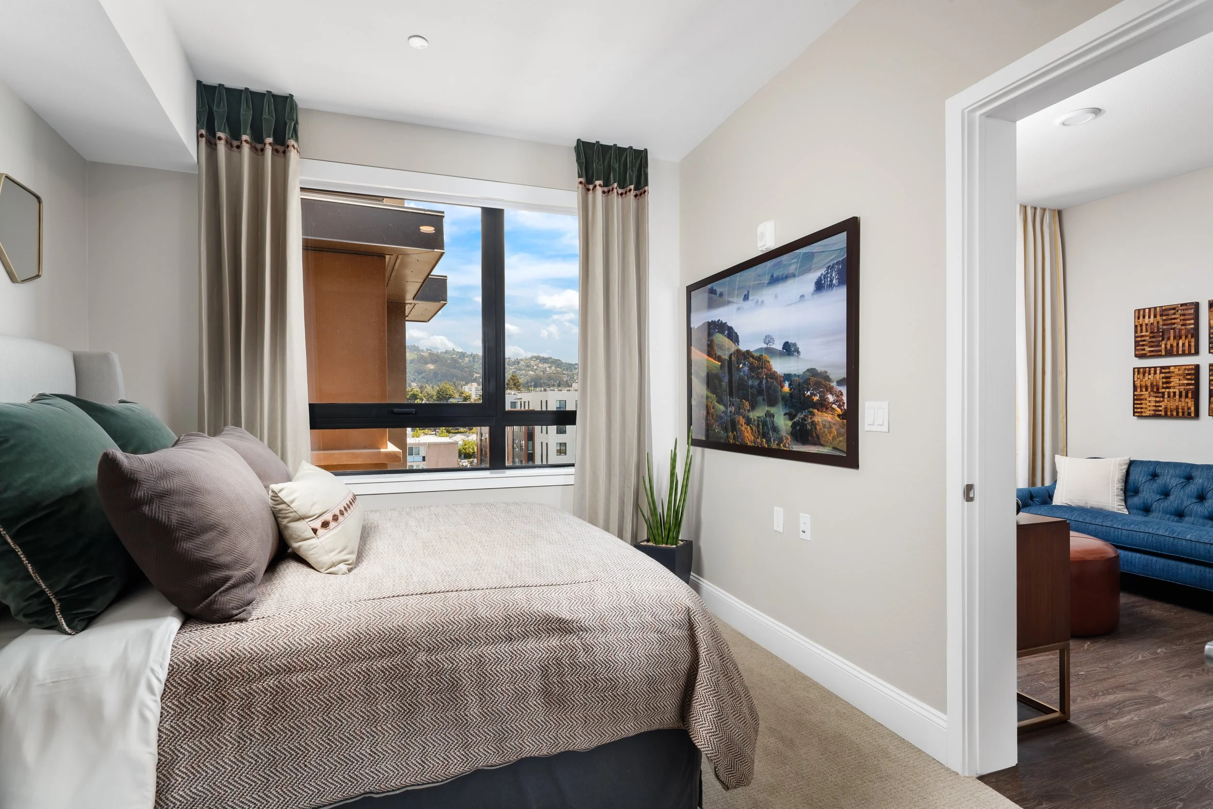 Bedroom with window view of city and hills, bed with multiple pillows, framed landscape photo on wall, potted plant near window, adjoining room with blue couch and wall art