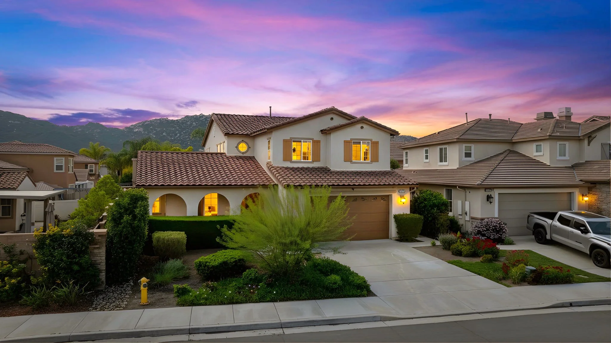 Suburban houses at sunset with mountains in the background, lit windows, and well-manicured lawns.