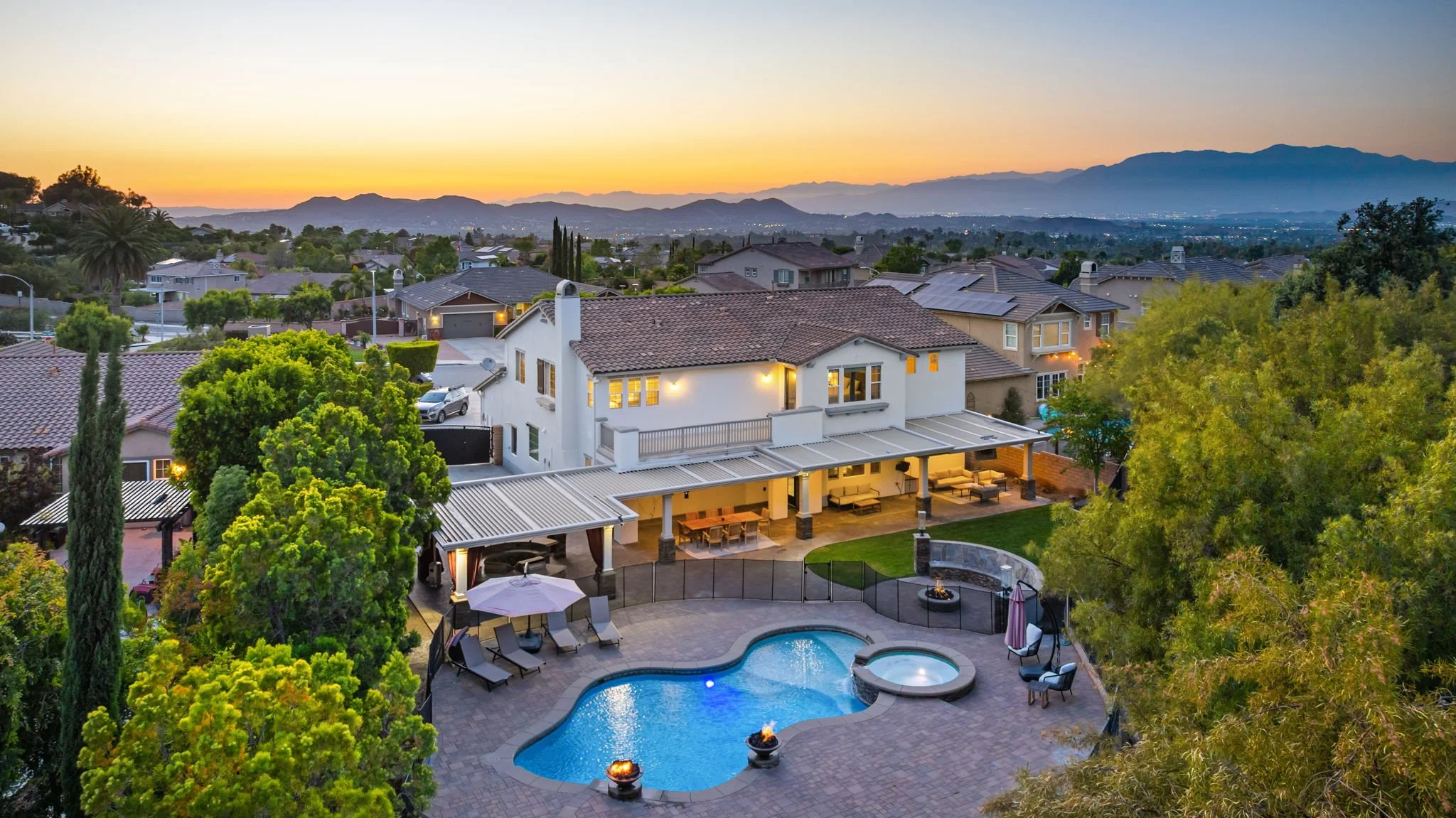 A backyard with a swimming pool, hot tub, outdoor seating, and fire pits, surrounded by trees and neighboring houses, with mountains in the distance during sunset.