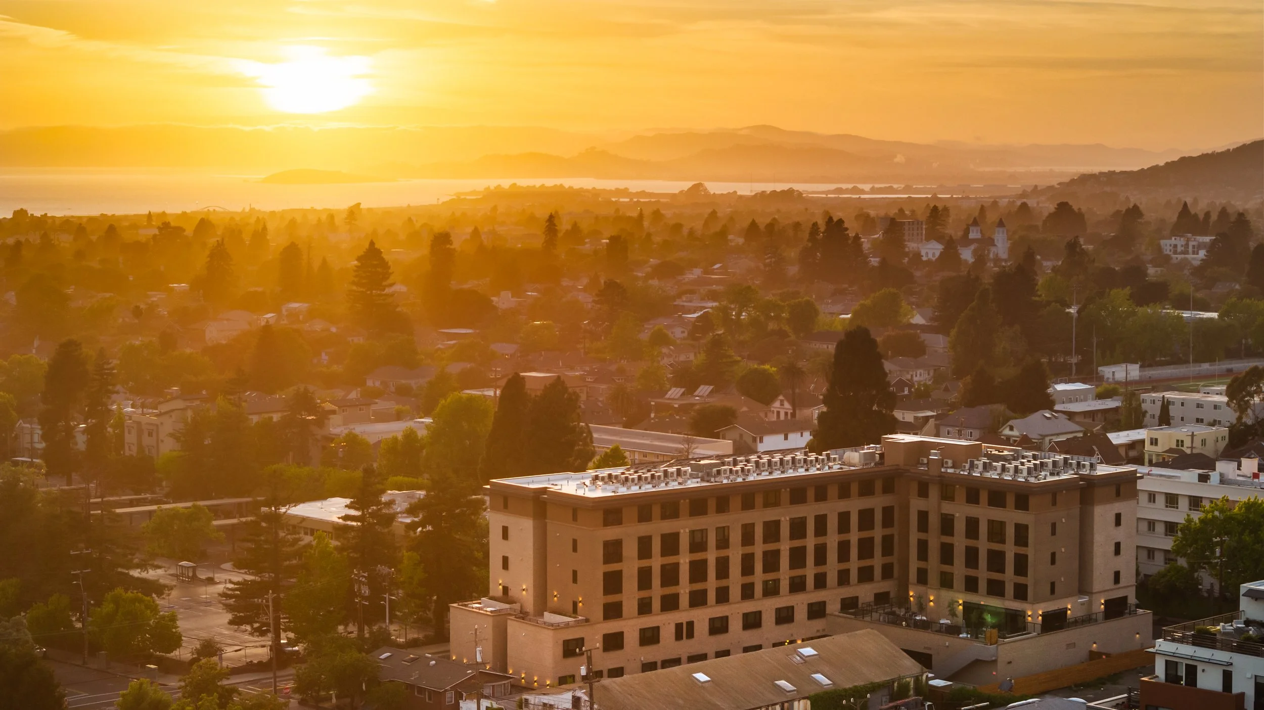 Sunset over a cityscape with a large building in the foreground, surrounded by trees and residential areas, with hills and water in the distance.