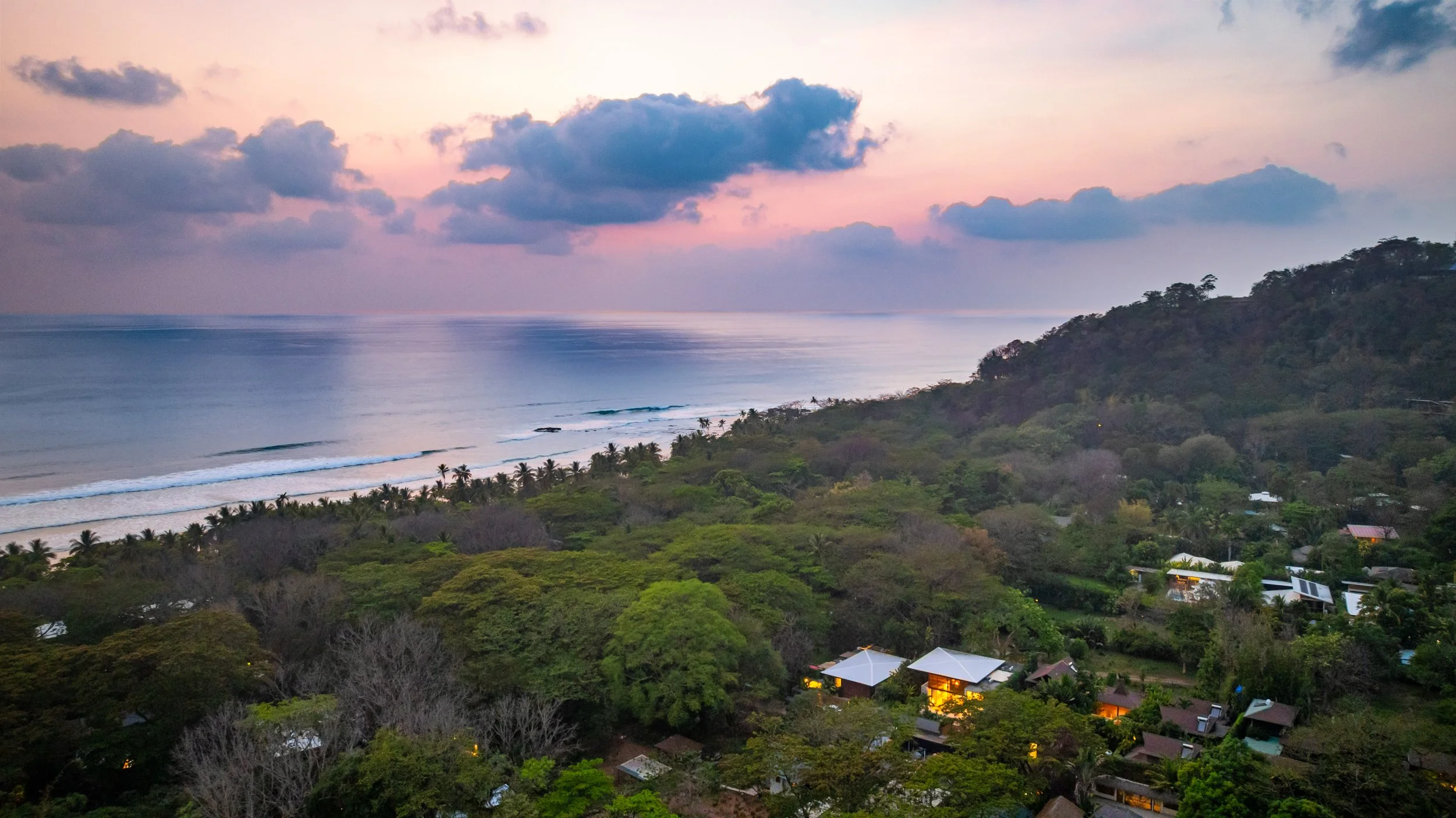 A scenic view of a coastline at sunset with a cloudy sky, calm ocean waves, a stretch of sandy beach with palm trees, and a lush green landscape with houses.
