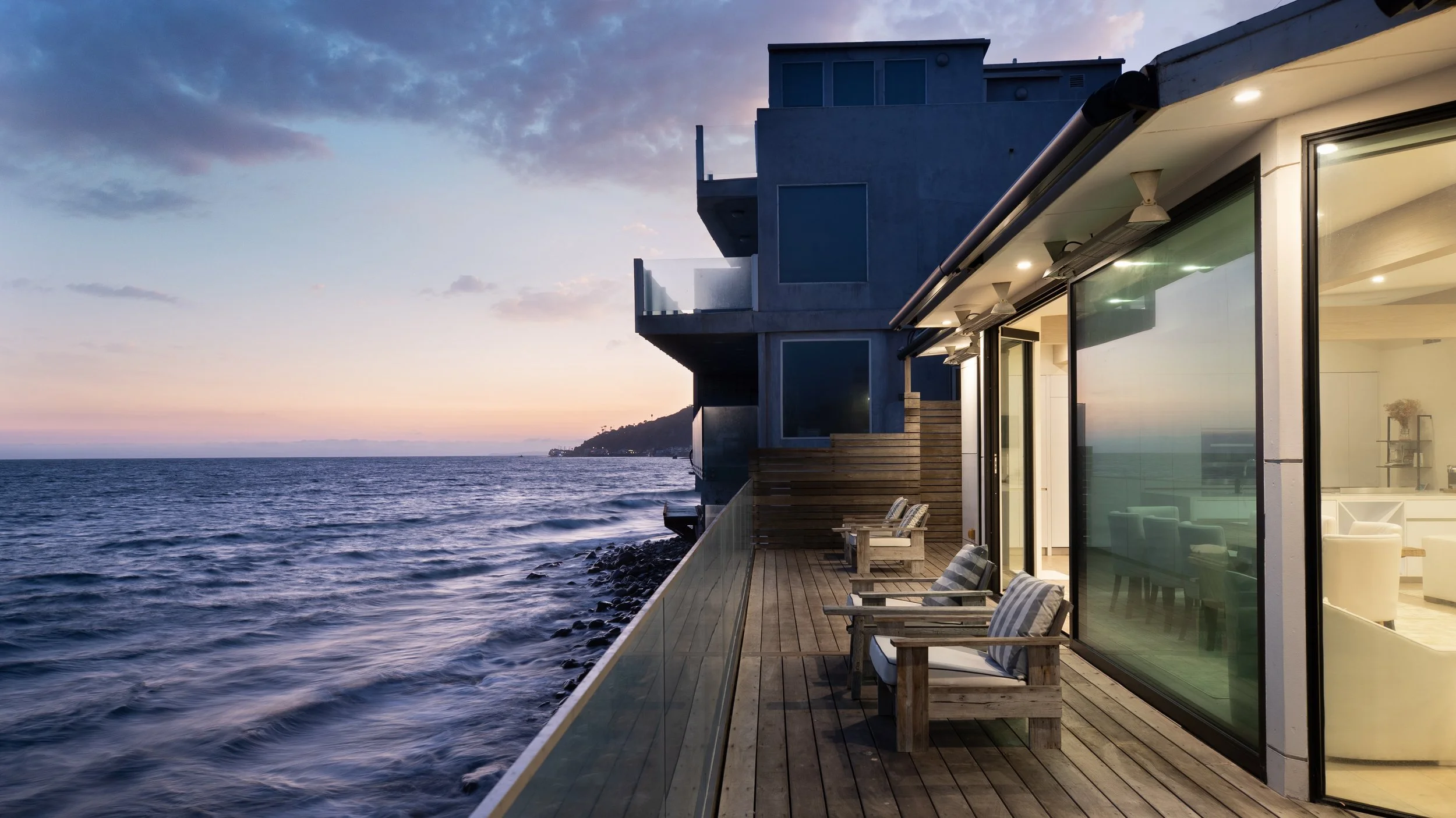 A sea view balcony of a modern house at sunset with wooden chairs and glass railing, overlooking the ocean and having an outdoor terrace with interior lights visible inside.