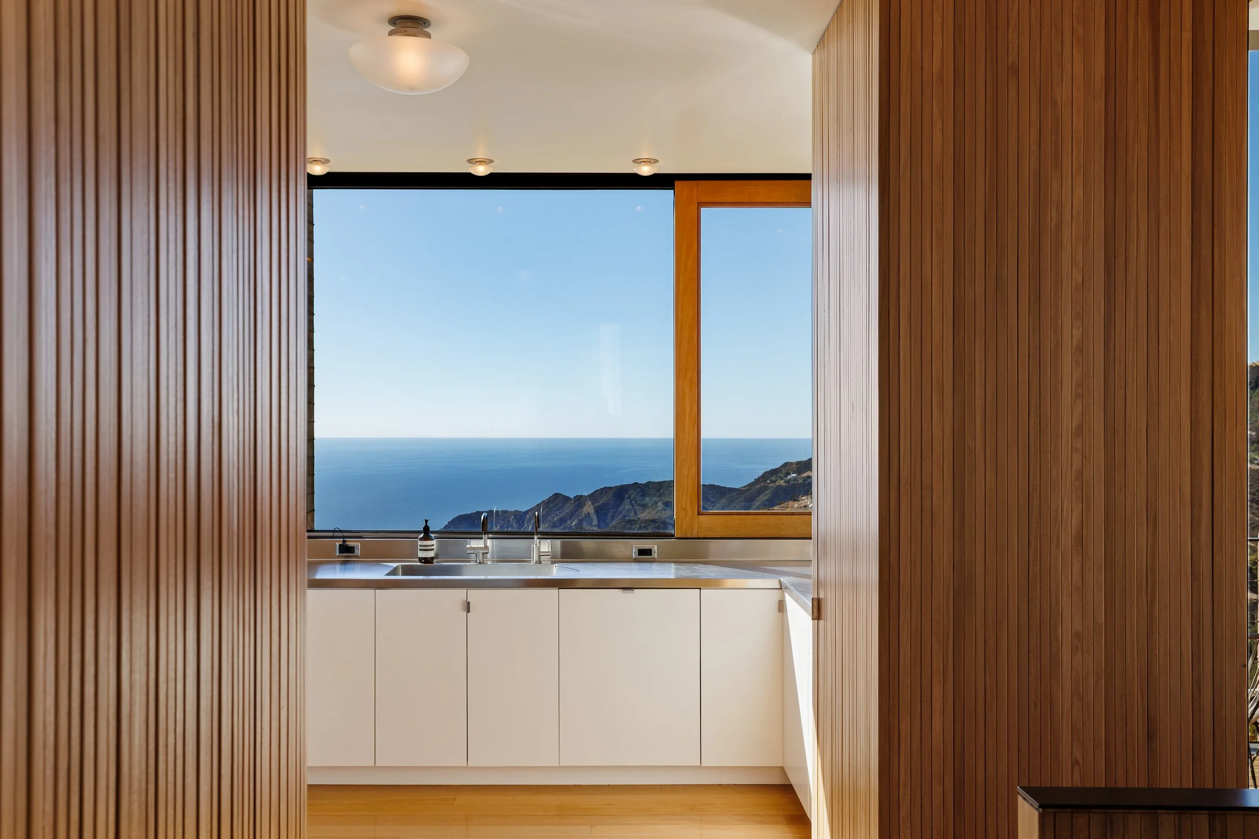 Kitchen with white cabinets, a stainless steel sink, and a large window showing a view of the ocean and distant mountains.
