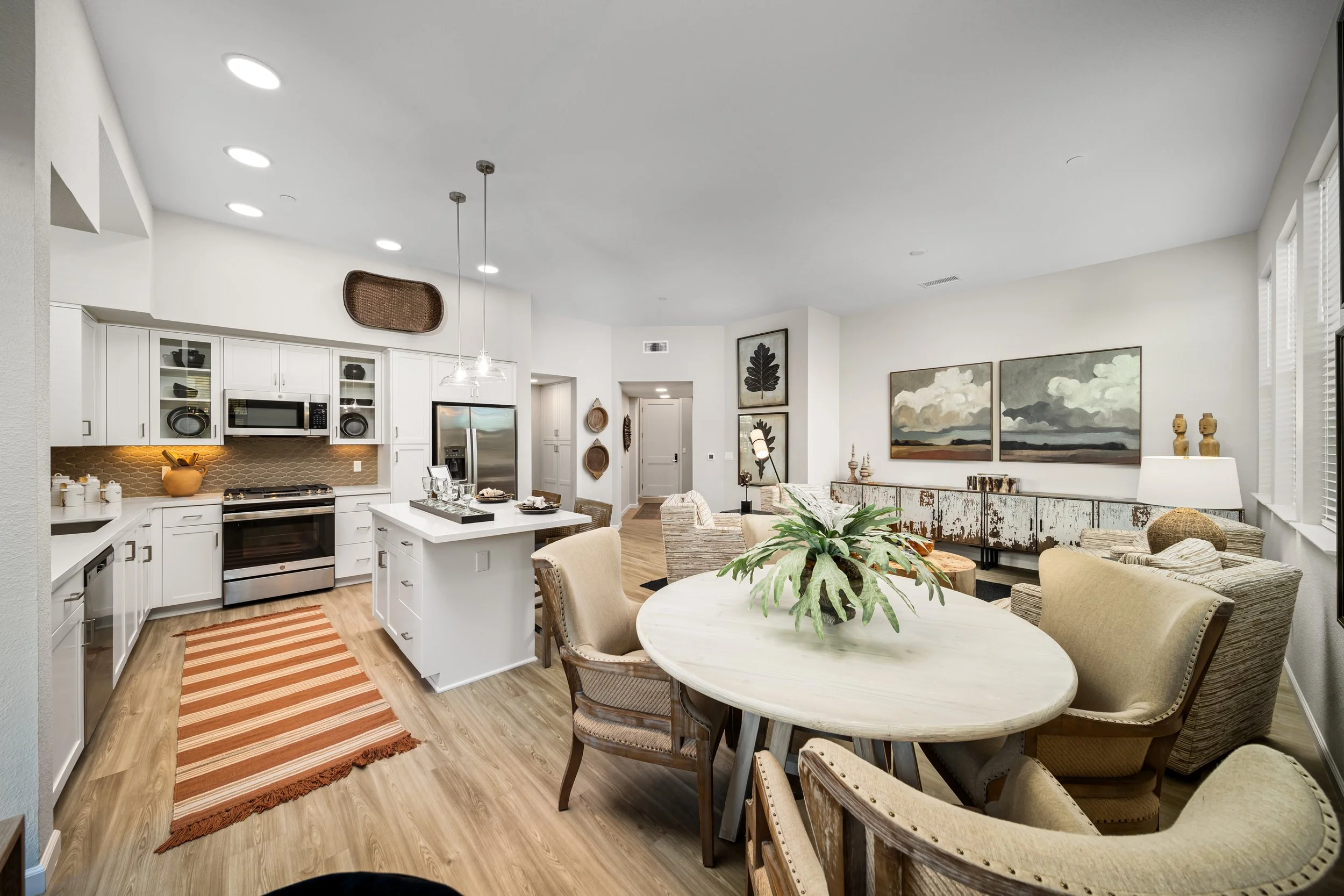 Open-concept kitchen and living room with white cabinets, stainless steel appliances, a round dining table with beige chairs, and artwork on the walls.