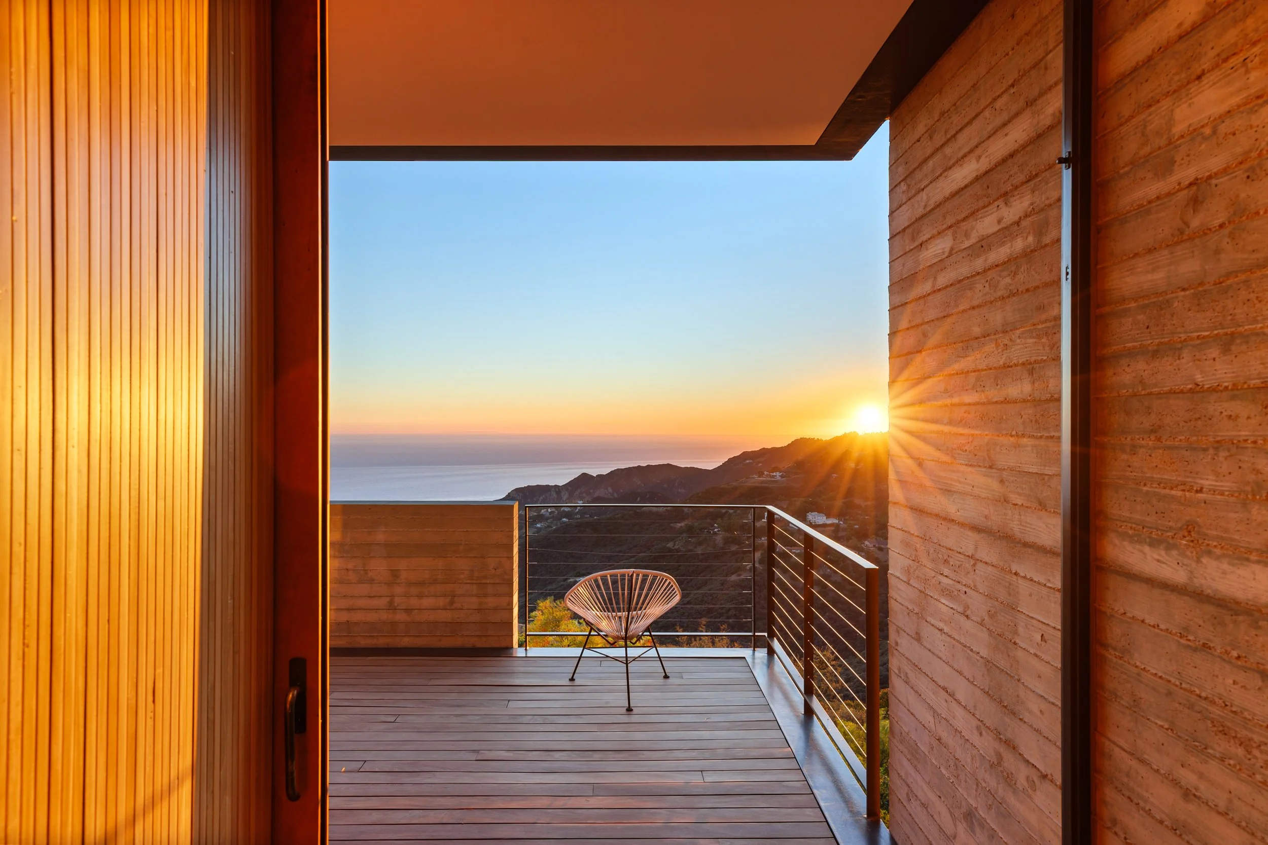 View of a sunset over a hillside and ocean from a wooden balcony with a single chair.