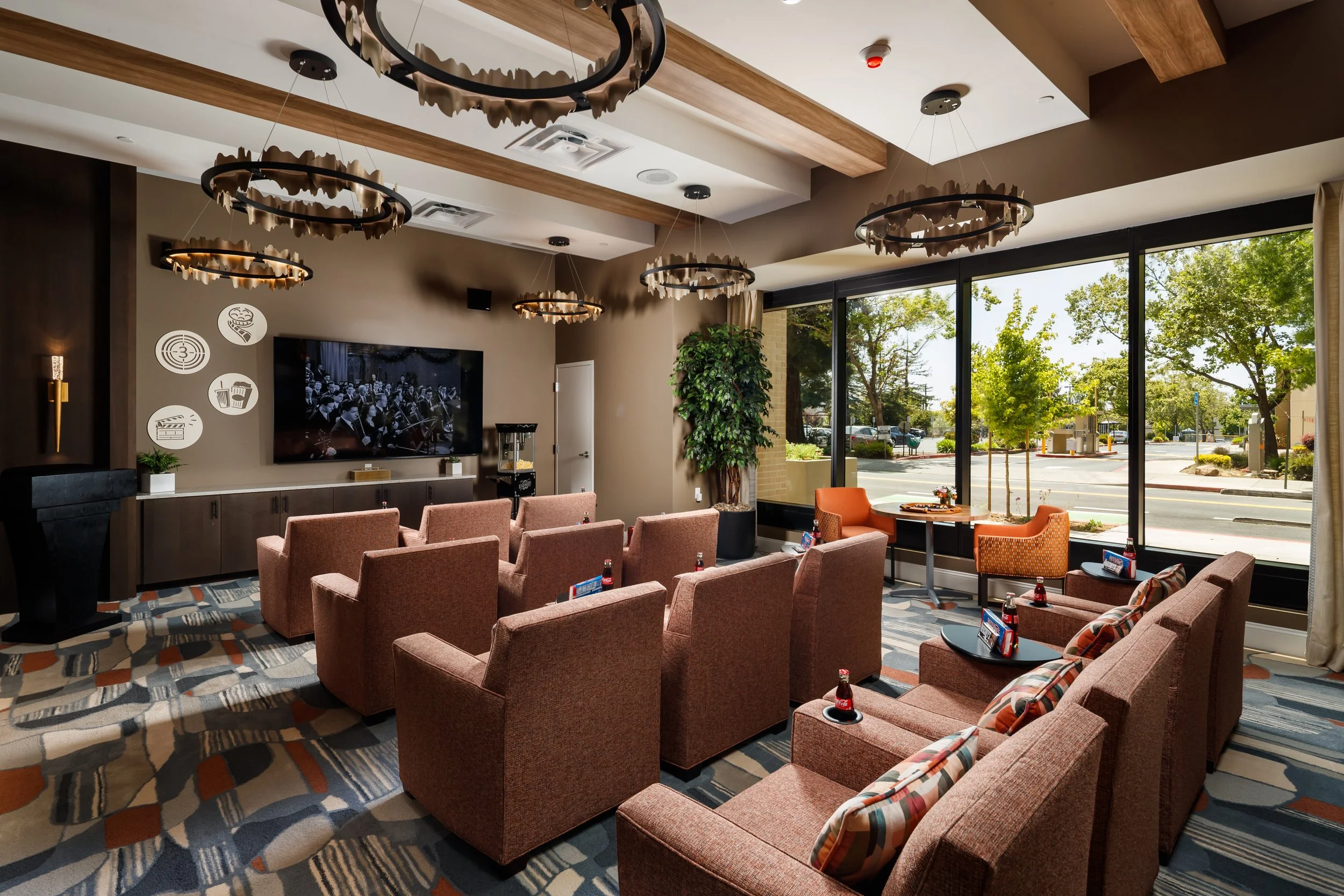 A cozy indoor lounge area with multiple rows of pink armchairs facing a large television, decorated with modern circular chandeliers, beside large windows revealing an outdoor parking lot and trees.
