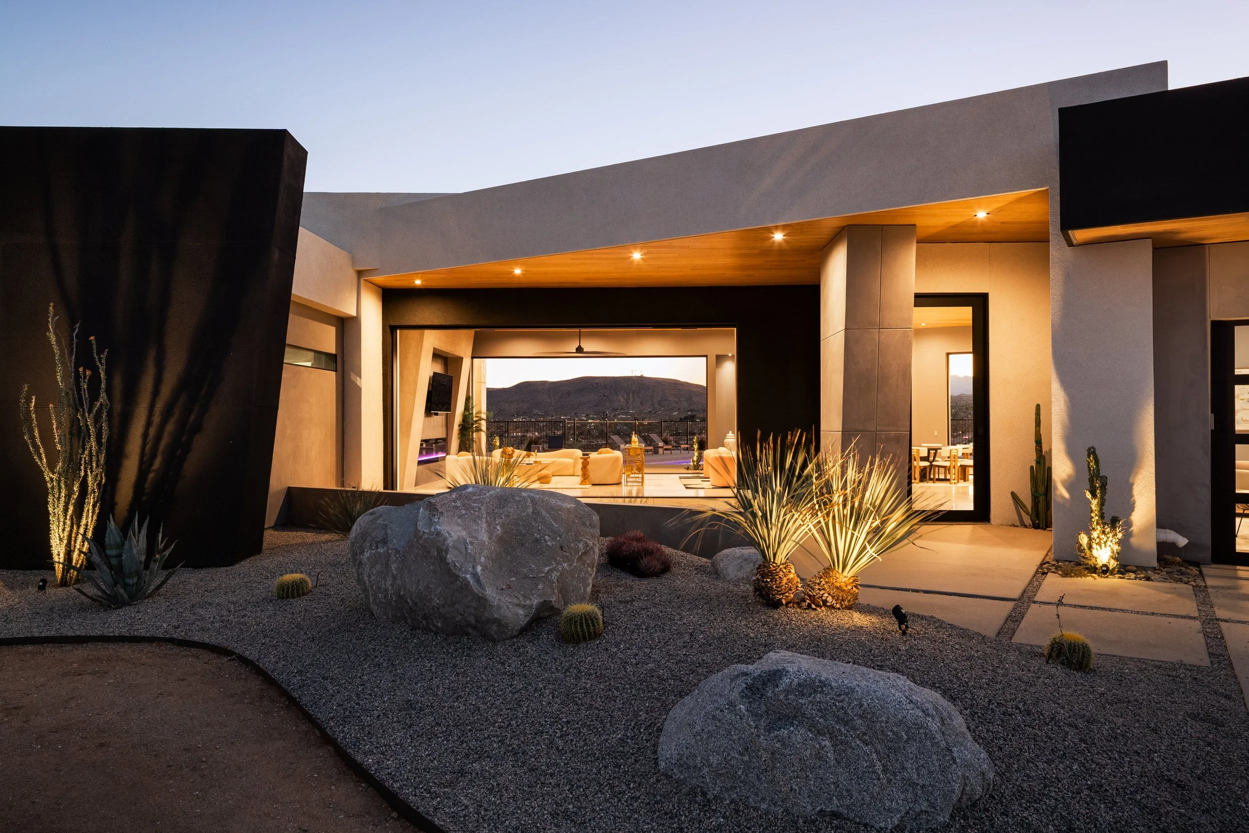 Modern house with desert landscaping, illuminated outdoor seating area, and hills in the background at sunset.