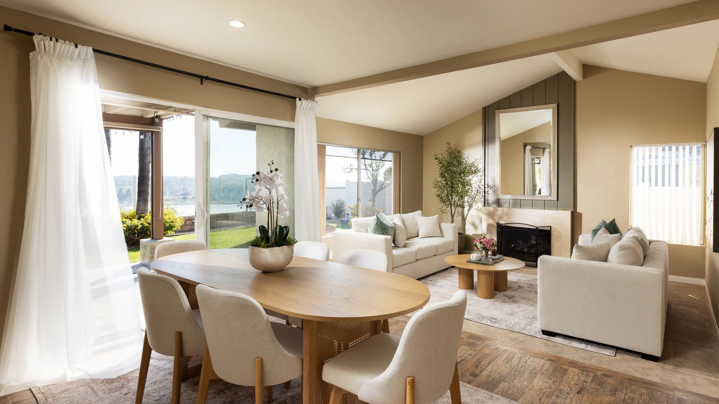 Bright living room with white sofas, a round wooden coffee table, and large windows showing a green outdoor area with trees and a lake. There are potted plants and a mirror above the fireplace.
