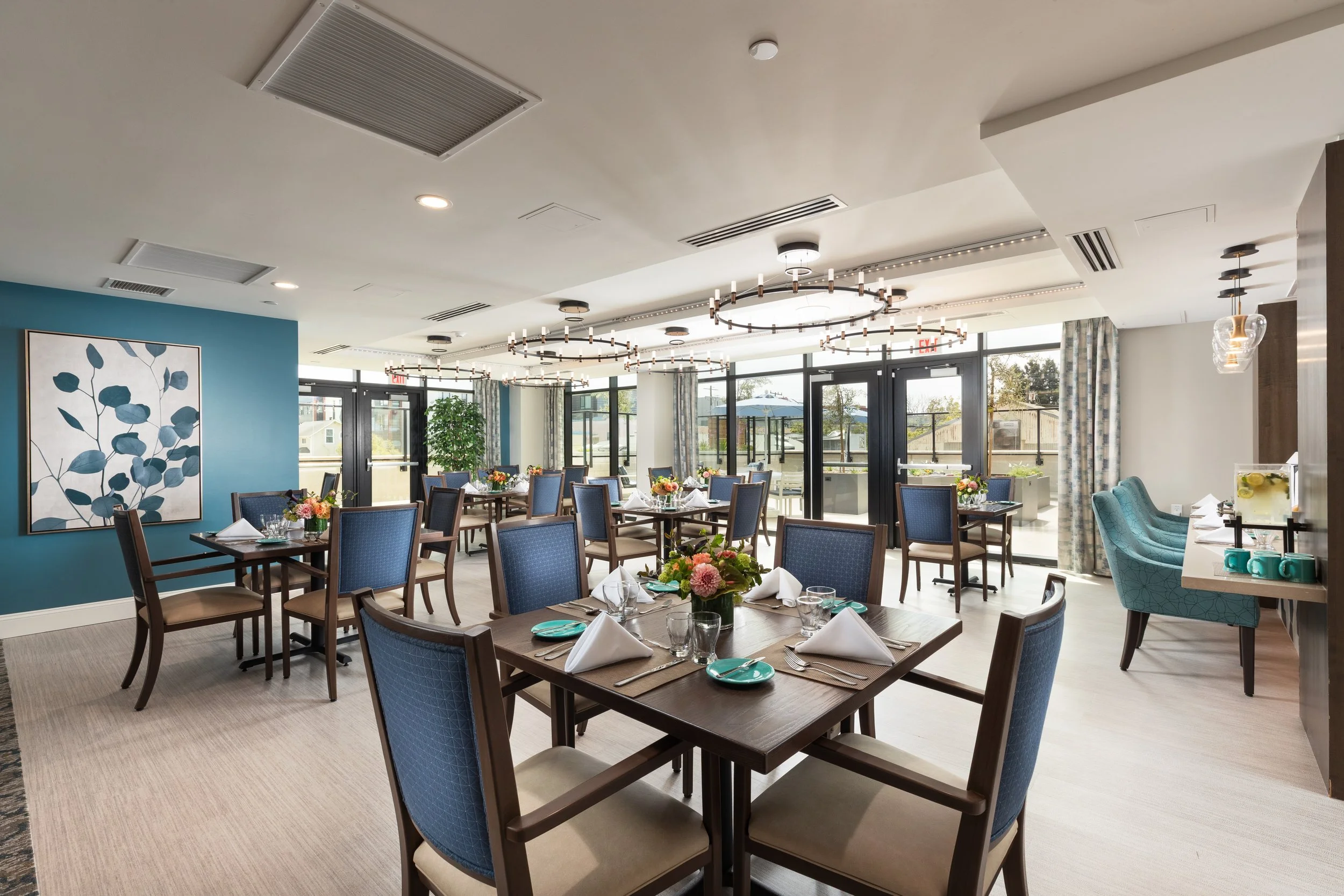 A modern restaurant or dining area with multiple tables set with white napkins, glassware, and floral centerpieces. The room features blue and beige chairs, a turquoise accent wall, large windows with curtains, and contemporary chandeliers. There is 