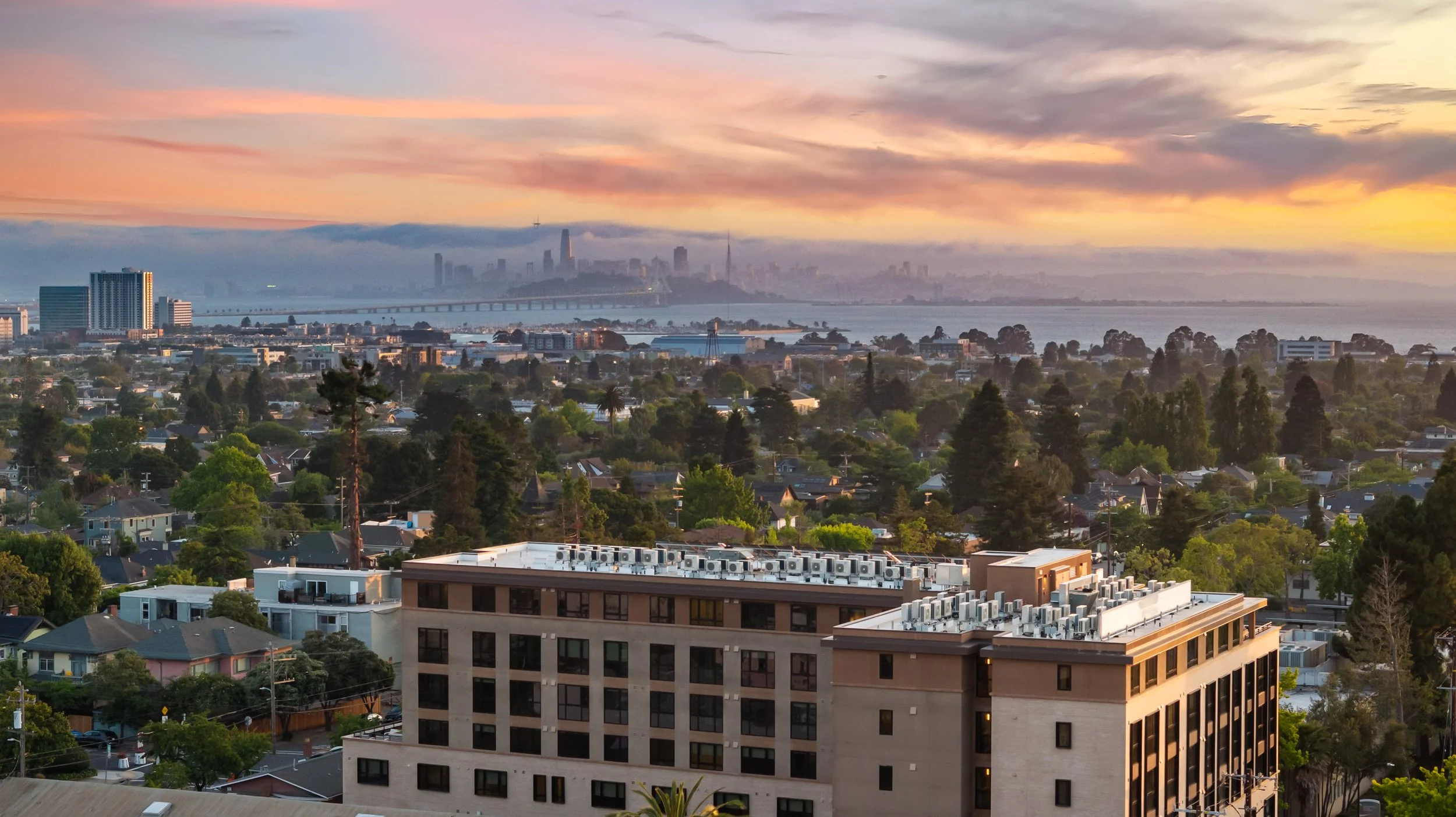 A city skyline at sunset with tall buildings, a bridge over water, and a foreground of residential houses and a mid-rise building with rooftop HVAC units.
