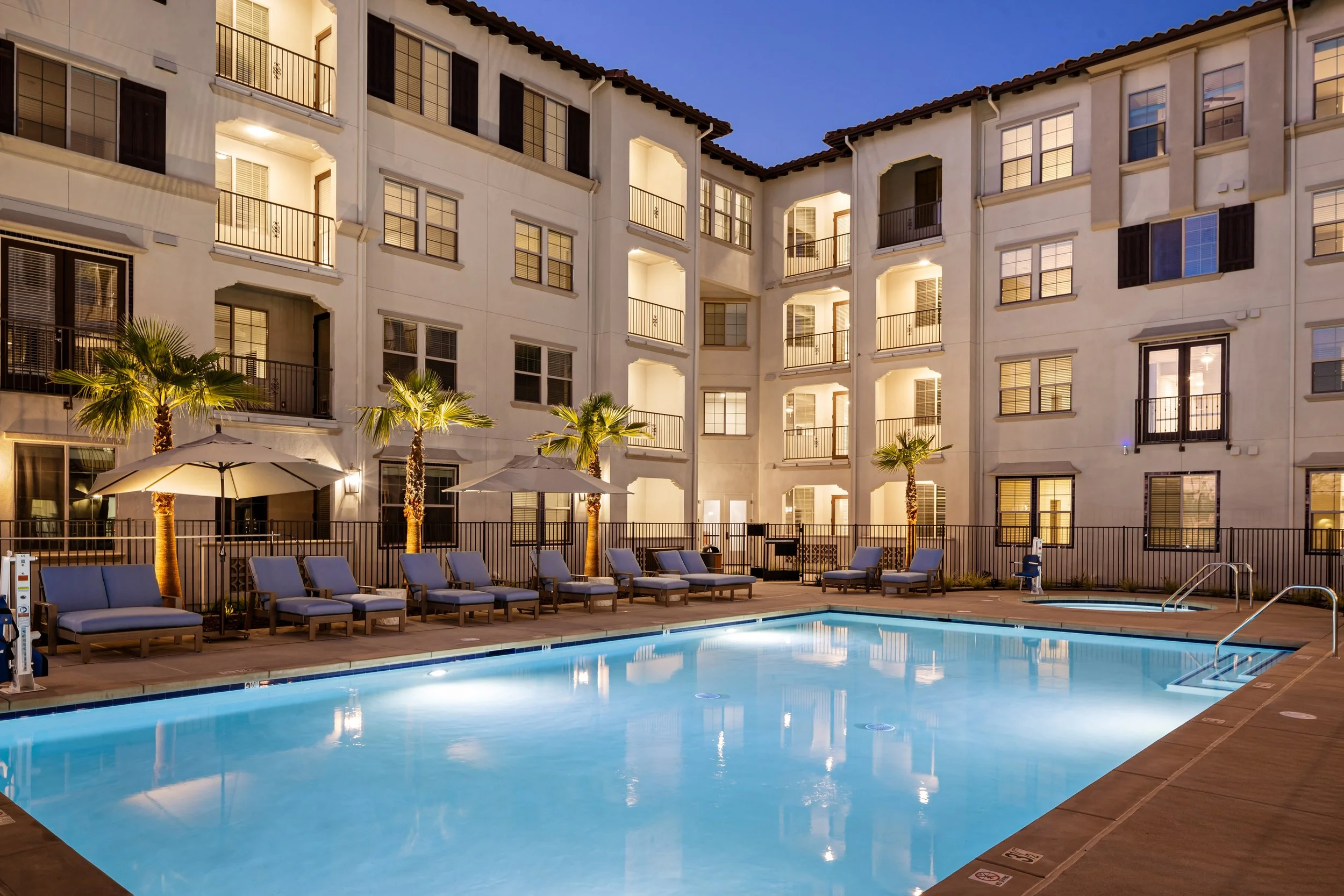 An outdoor swimming pool at dusk with lounge chairs, umbrellas, and small palm trees, surrounded by a fenced area and multi-story apartment buildings with balconies, illuminated from inside.