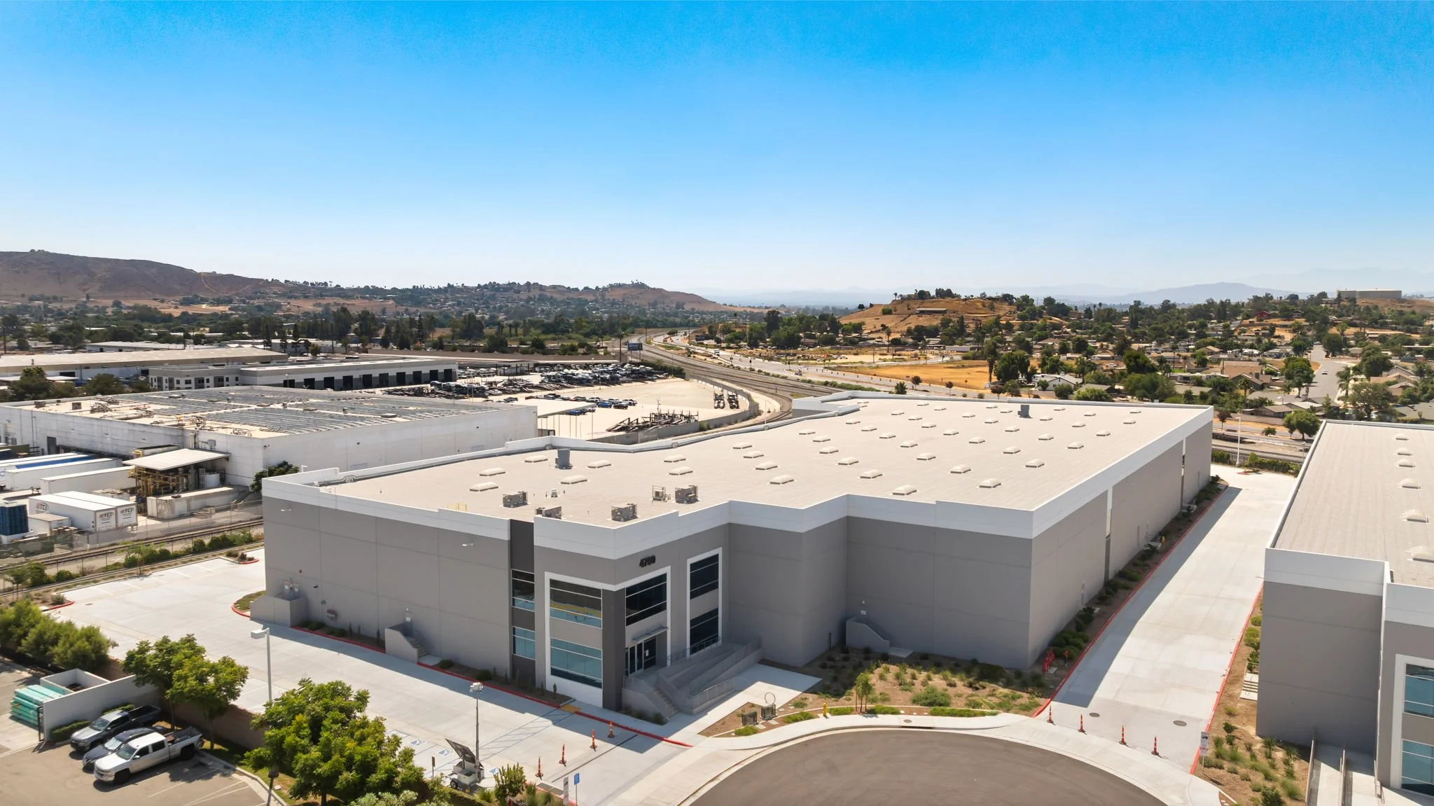 An aerial view of a large white commercial or industrial building with a parking lot and adjacent roads, overlooking a hilly landscape and a suburban area under clear blue skies.